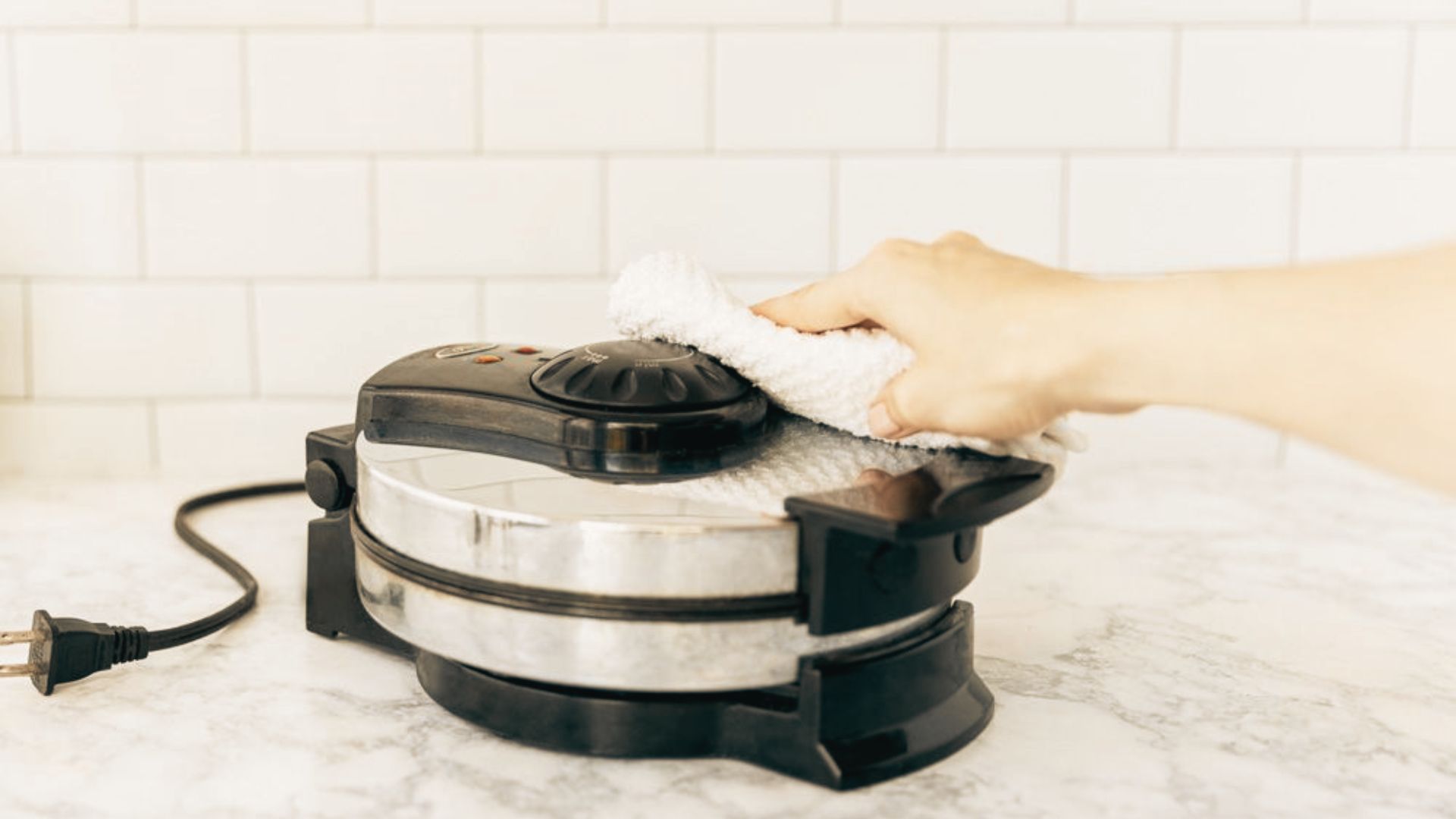 A hand wipes the exterior of a closed, unplugged waffle maker with a white cloth on a marble counter.