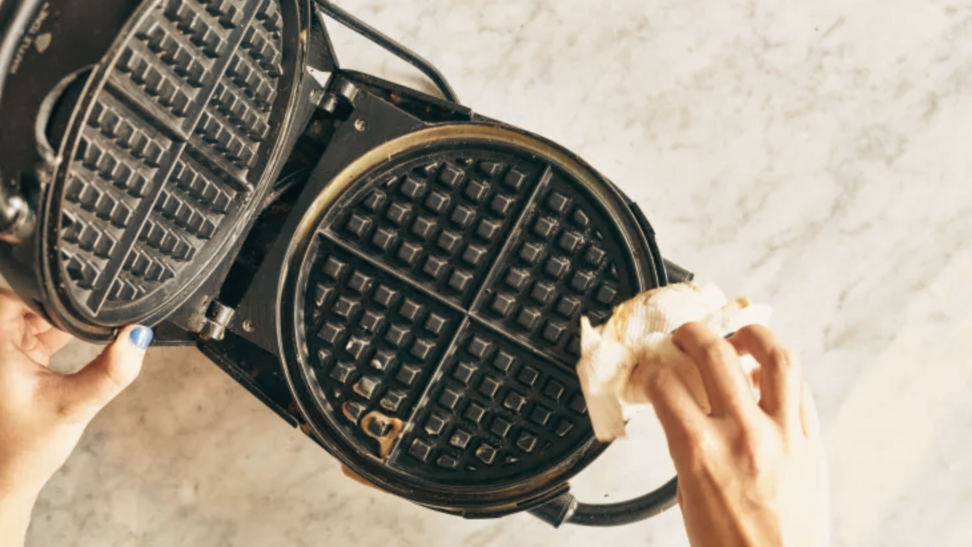 Top-down view of hands wiping batter residue from an open waffle maker with a paper towel.