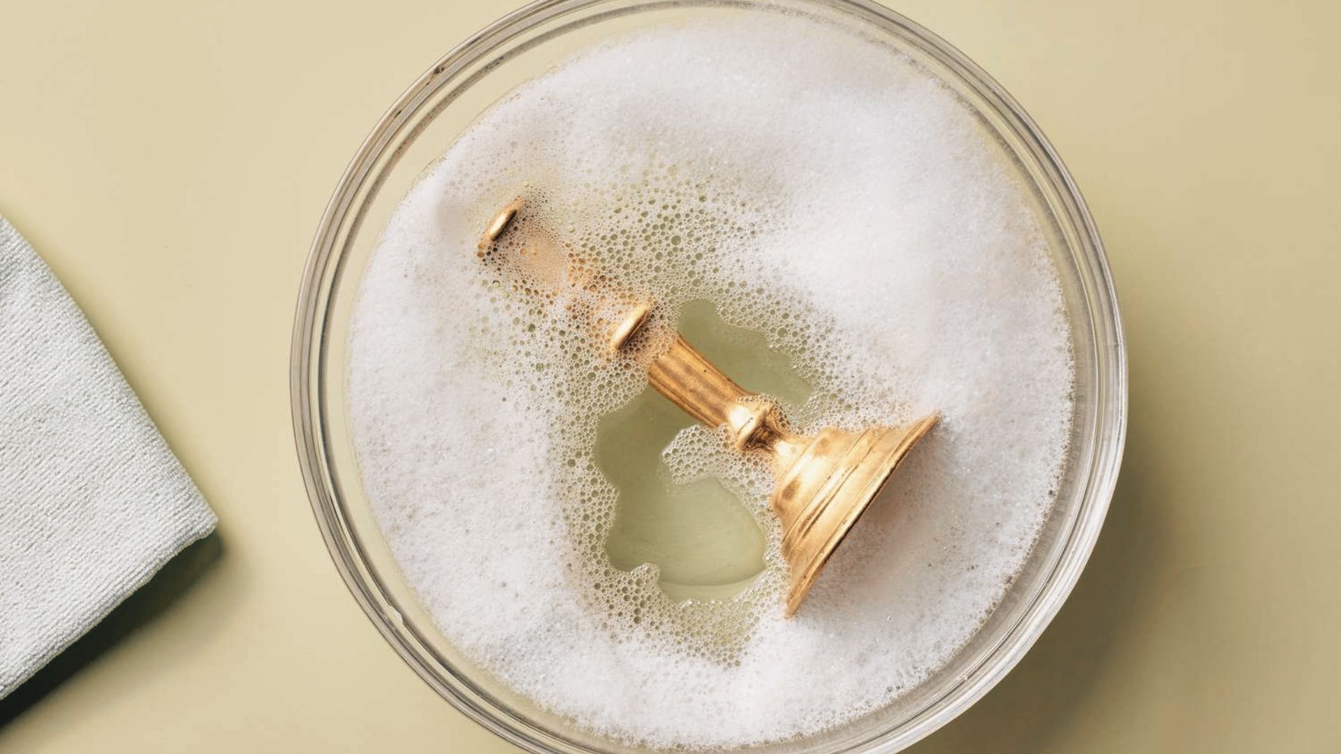 Brass candle holder soaking in soapy water inside a glass bowl next to a soft cleaning cloth.