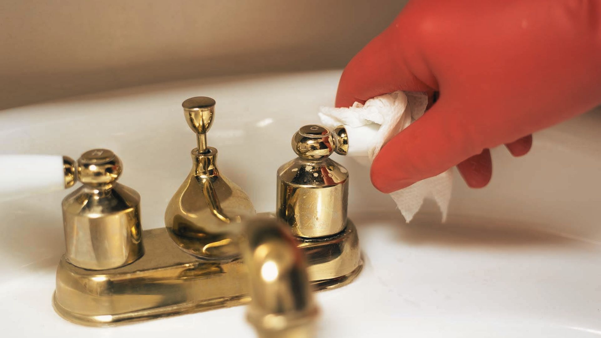 Red-gloved hand wiping brass bathroom faucet with white cloth to clean water spots and tarnish.