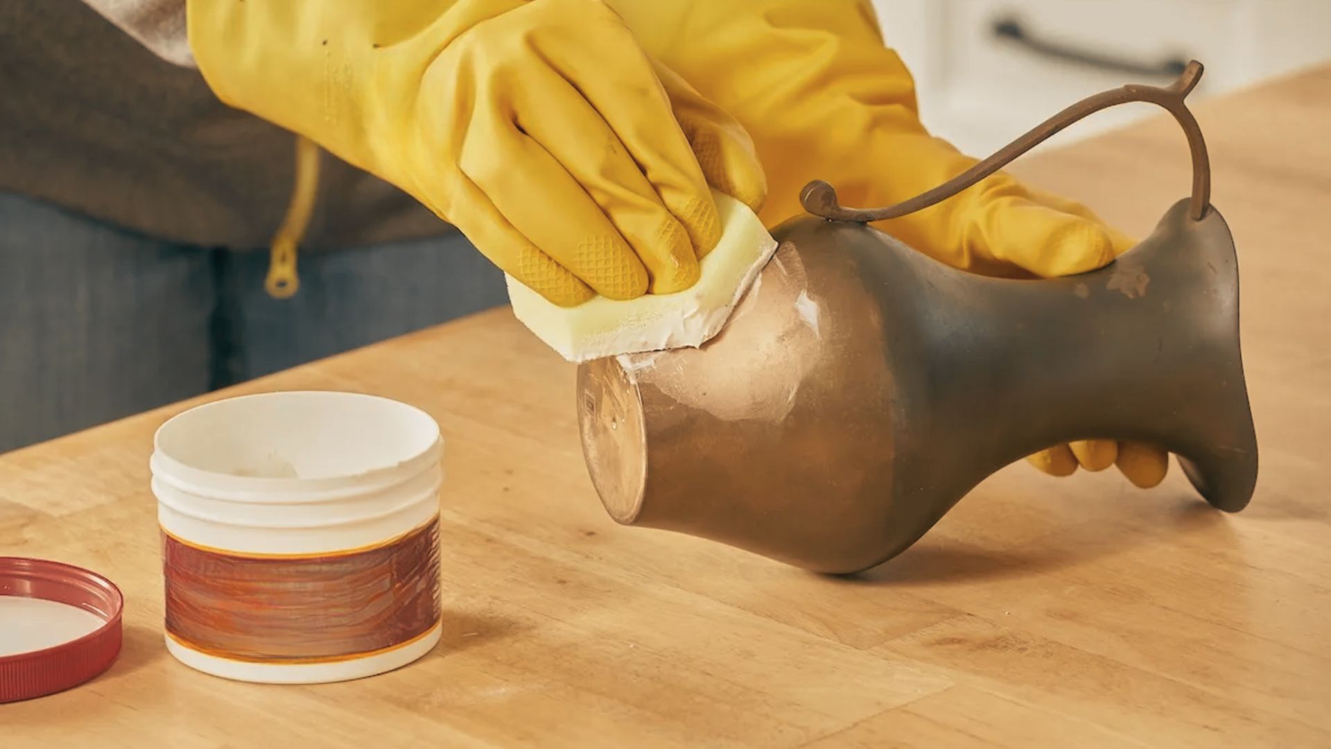 Hands in yellow gloves scrubbing a brass vase with paste using a sponge to lift heavy tarnish.