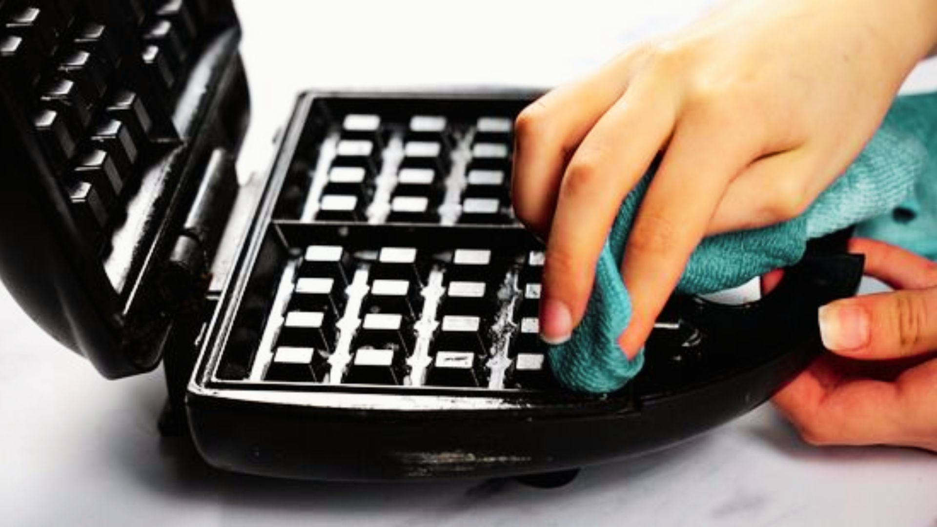 Hands use a teal microfiber cloth to wipe cleaning solution from the grids of an open waffle maker.