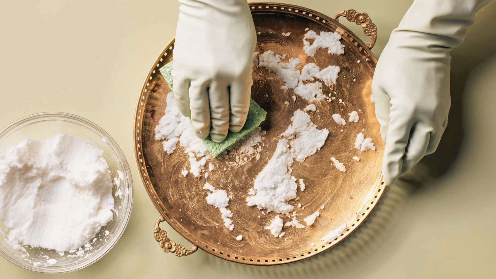 Gloved hands applying white cleaning paste to an ornate brass tray with sponge to remove tarnish.