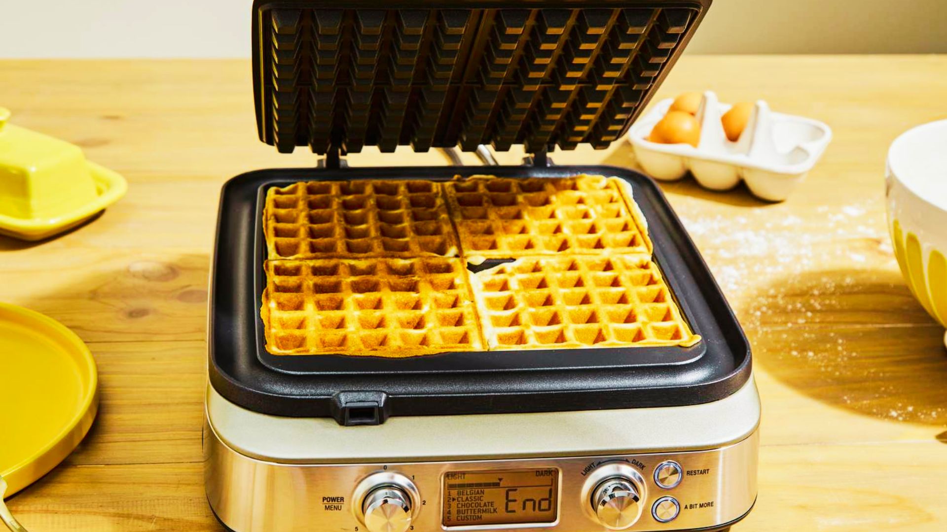 A Heavy-Build waffle maker open with four golden waffles inside, surrounded by eggs and butter on a wooden counter.