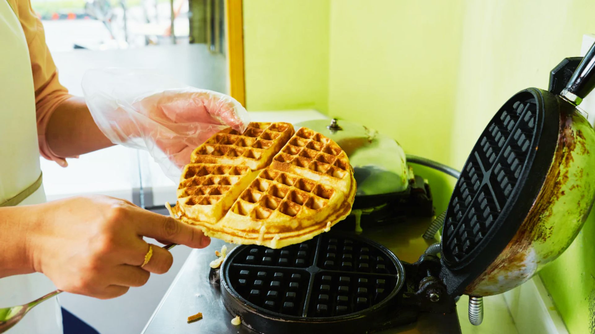 A person wearing gloves lifts a large golden waffle from a round waffle iron with visible batter residue on the grates.