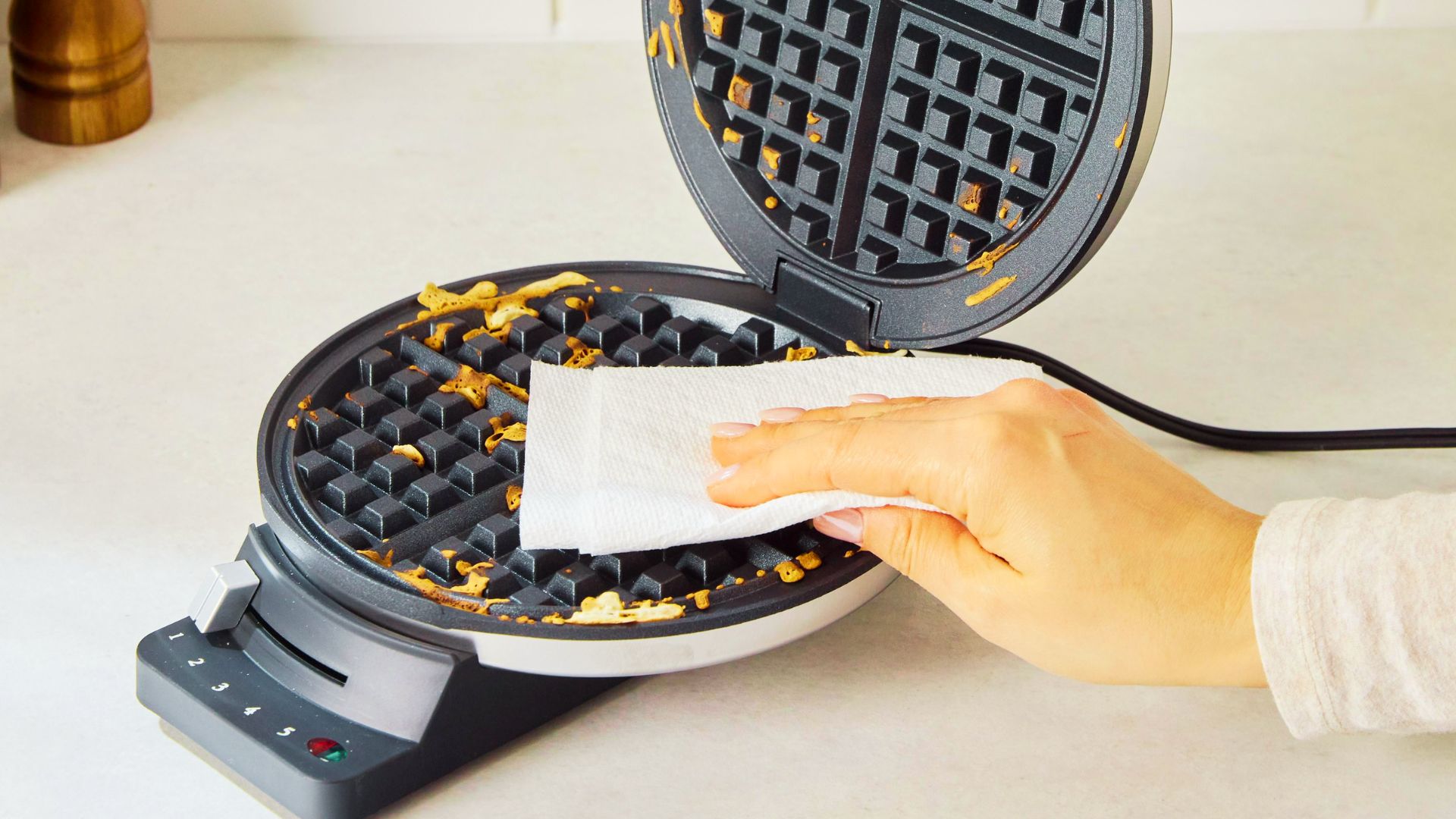 A hand uses a white paper towel to wipe batter crumbs from the grids of an open, unplugged waffle maker.