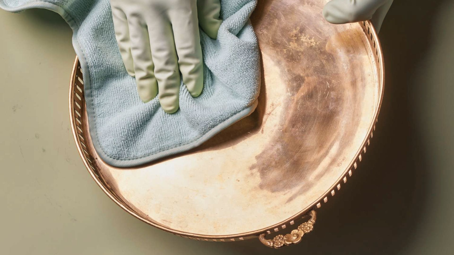Gloved hand buffing a brass tray with a blue microfiber cloth to restore shine and remove stains.