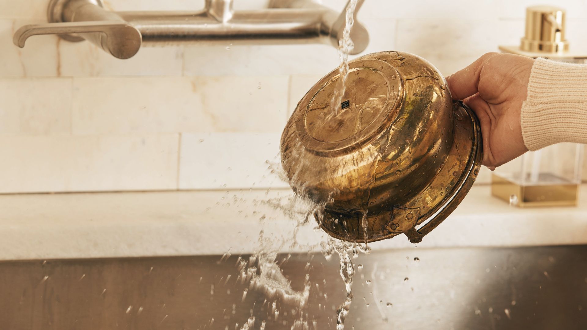 Hand rinsing tarnished brass bowl under running tap water to remove loose dirt and grime.