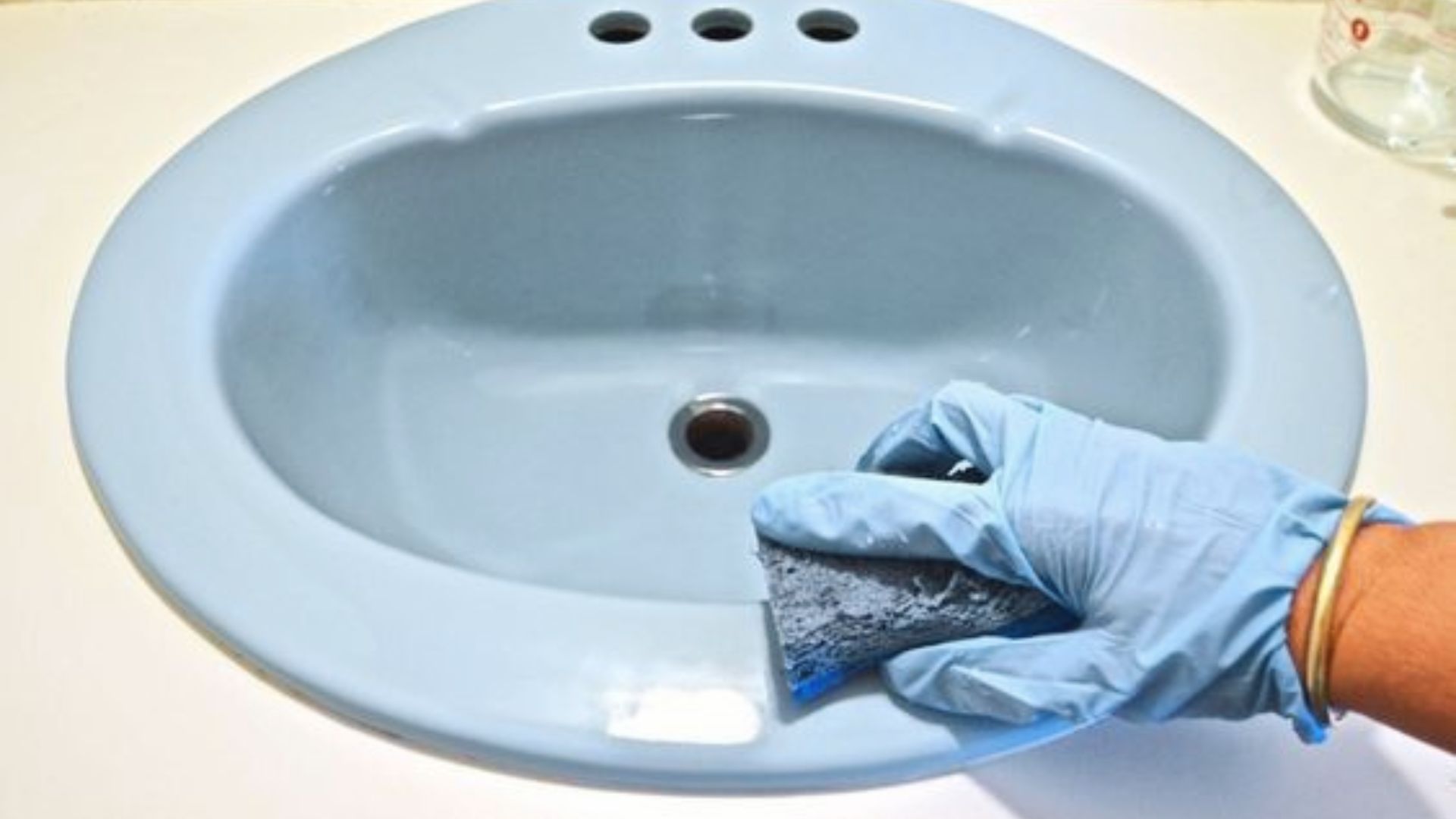 Gloved hand sanding a light blue bathroom sink basin as prep step for vanity refinishing project.