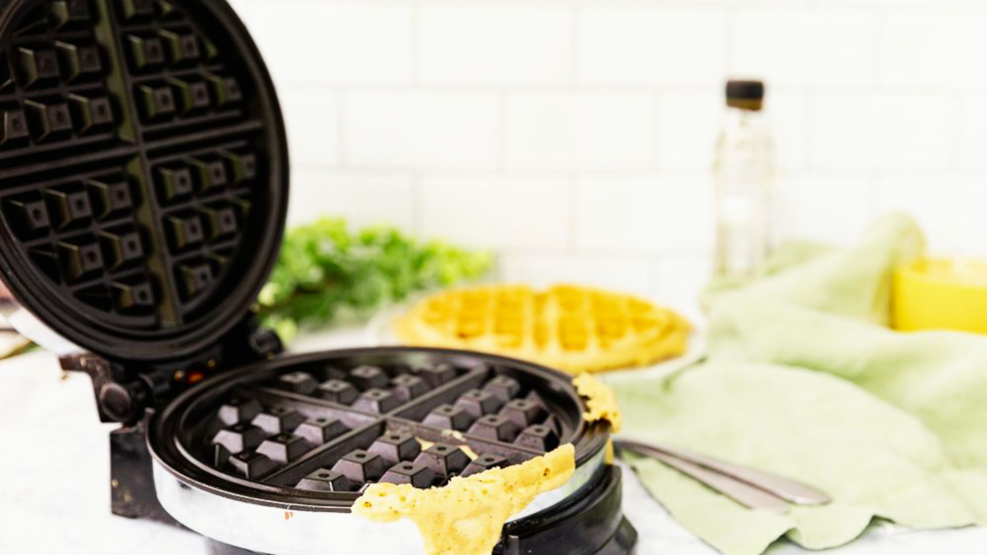 An open waffle maker with batter spilling over the edge sits next to a cooked waffle and green cloth.