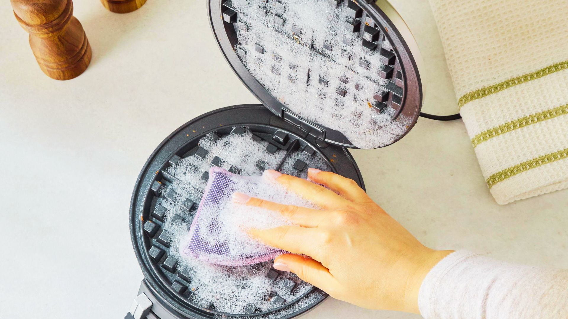 An open waffle maker with soapy suds covering both grids as hand scrubs with a purple sponge.