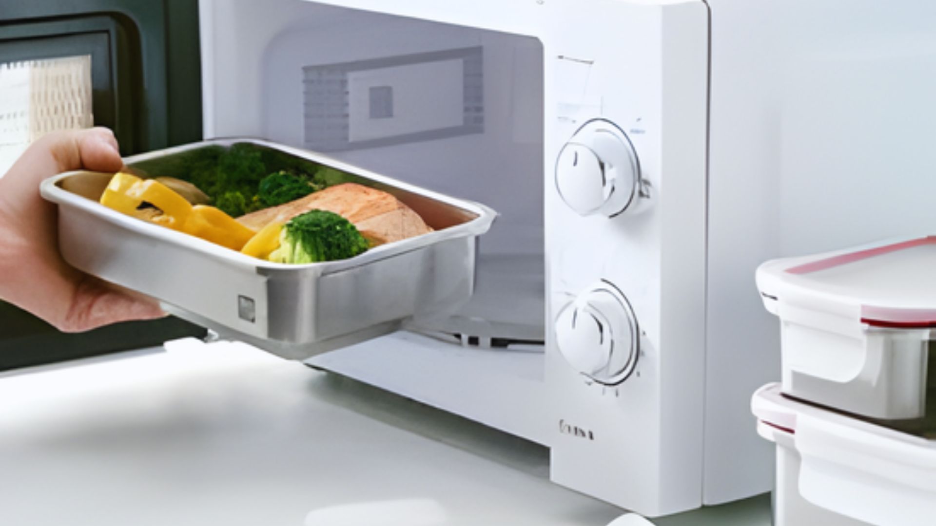 A person stands holding a tray of food in front of a microwave, preparing to heat the meal.