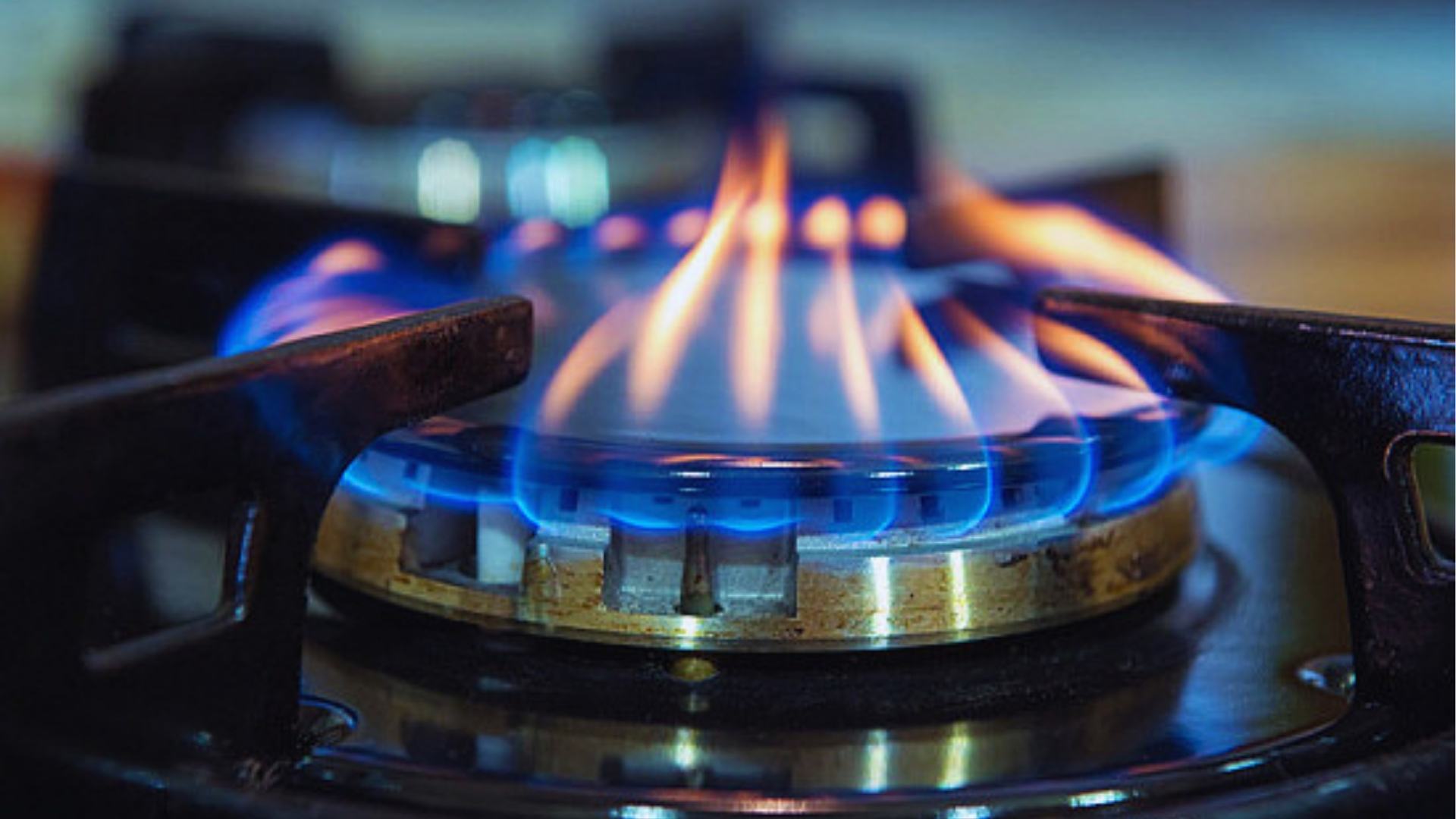 Close-up of bright blue gas flame burning on stove burner with black cast iron grates and blurred kitchen background.