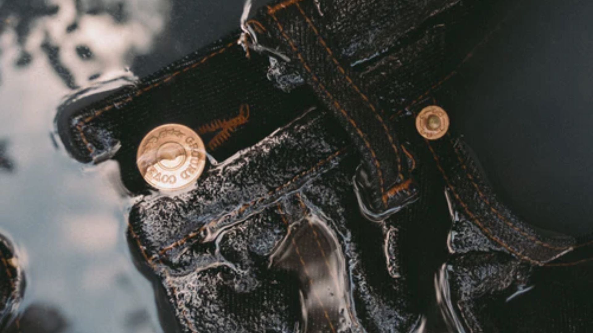 Close-up of wet black jeans with orange stitching and brass buttons submerged in water.