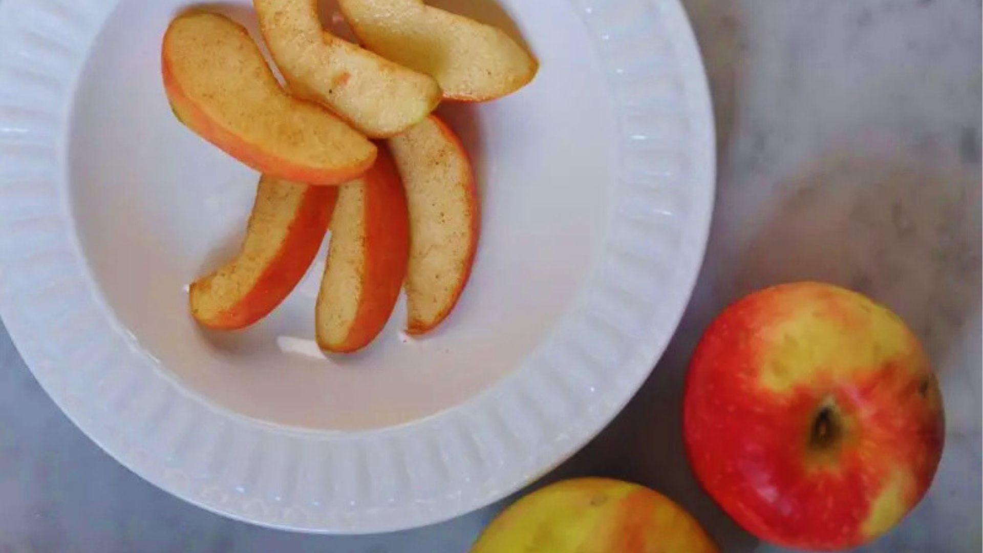 Apple slices dusted on white plate with two whole red apples nearby on a gray marble countertop.