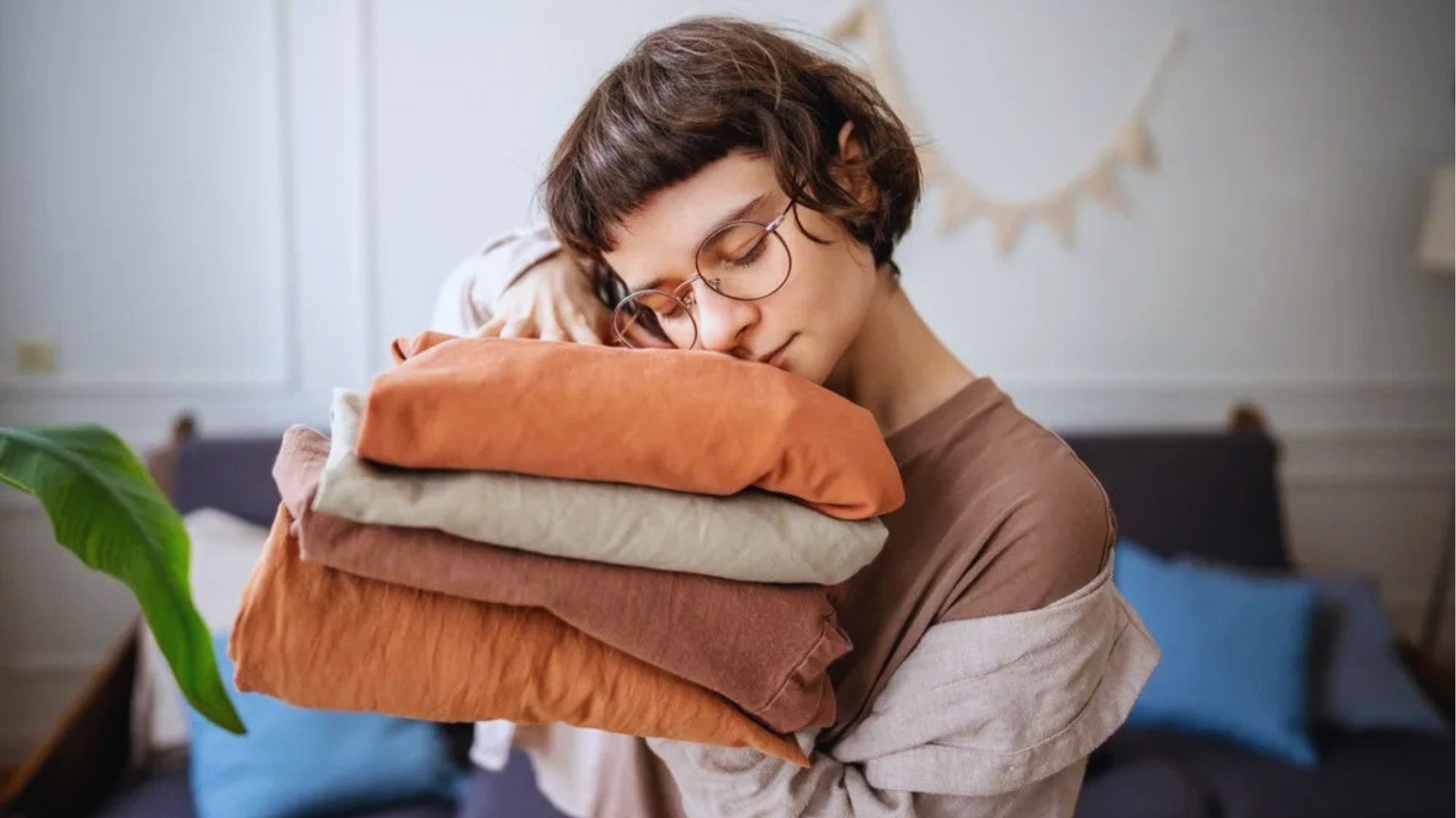 Women with short hair and glasses hugging a stack of folded clean laundry in earth tones against a cozy living room background.