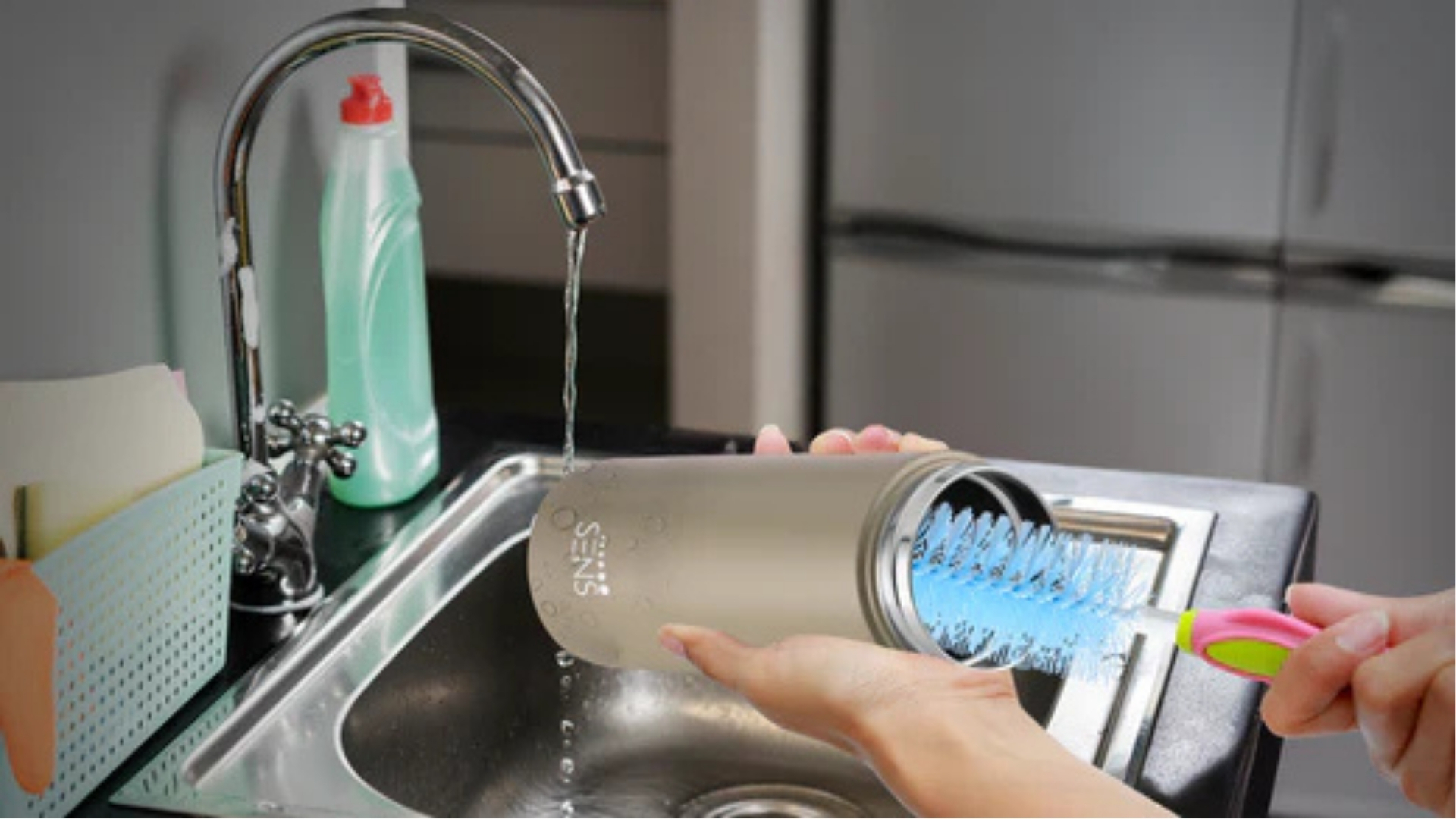 Person hand-washing beige stainless steel tumbler with blue bottle brush under running faucet in kitchen sink.