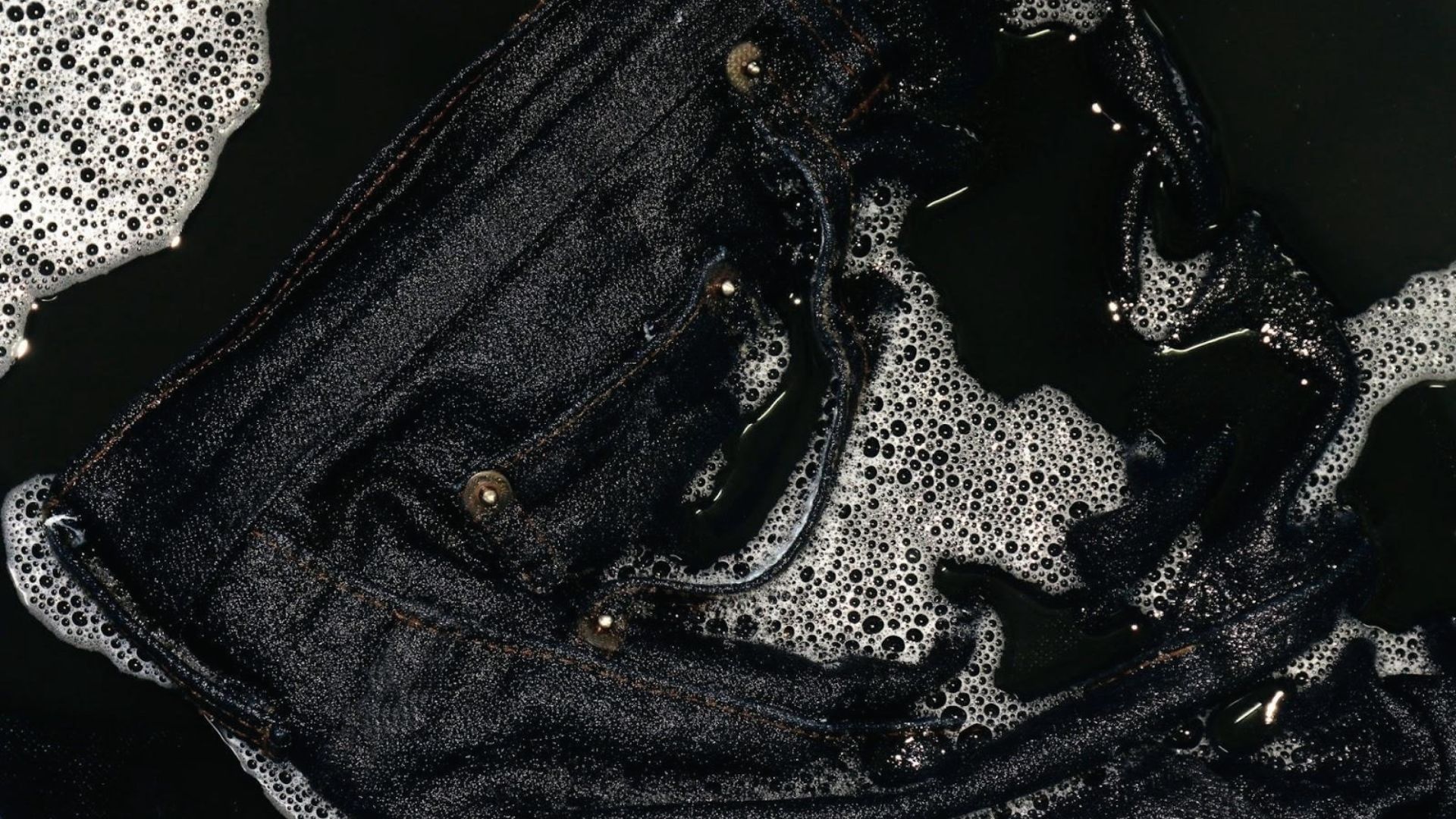 Close-up of black jeans submerged in dark water surrounded by soap bubbles.