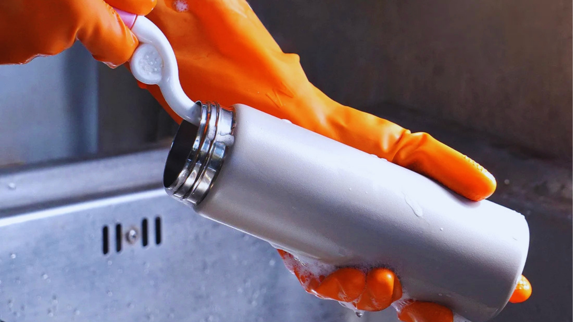 Person wearing orange rubber gloves scrubbing inside matte white stainless steel bottle with curved white bottle brush over sink.
