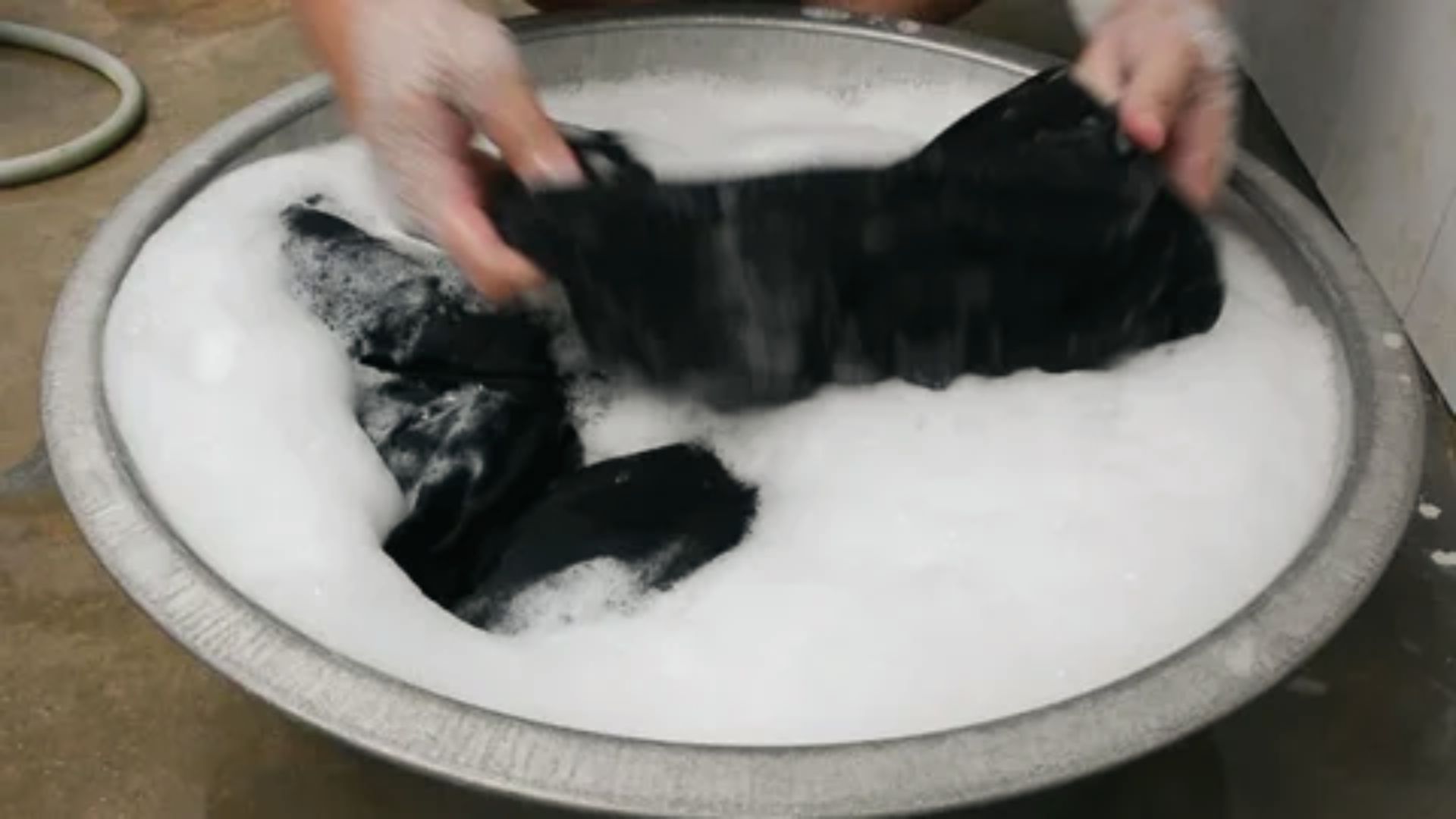 Hands gently agitate black jeans in a metal tub filled with soapy water.