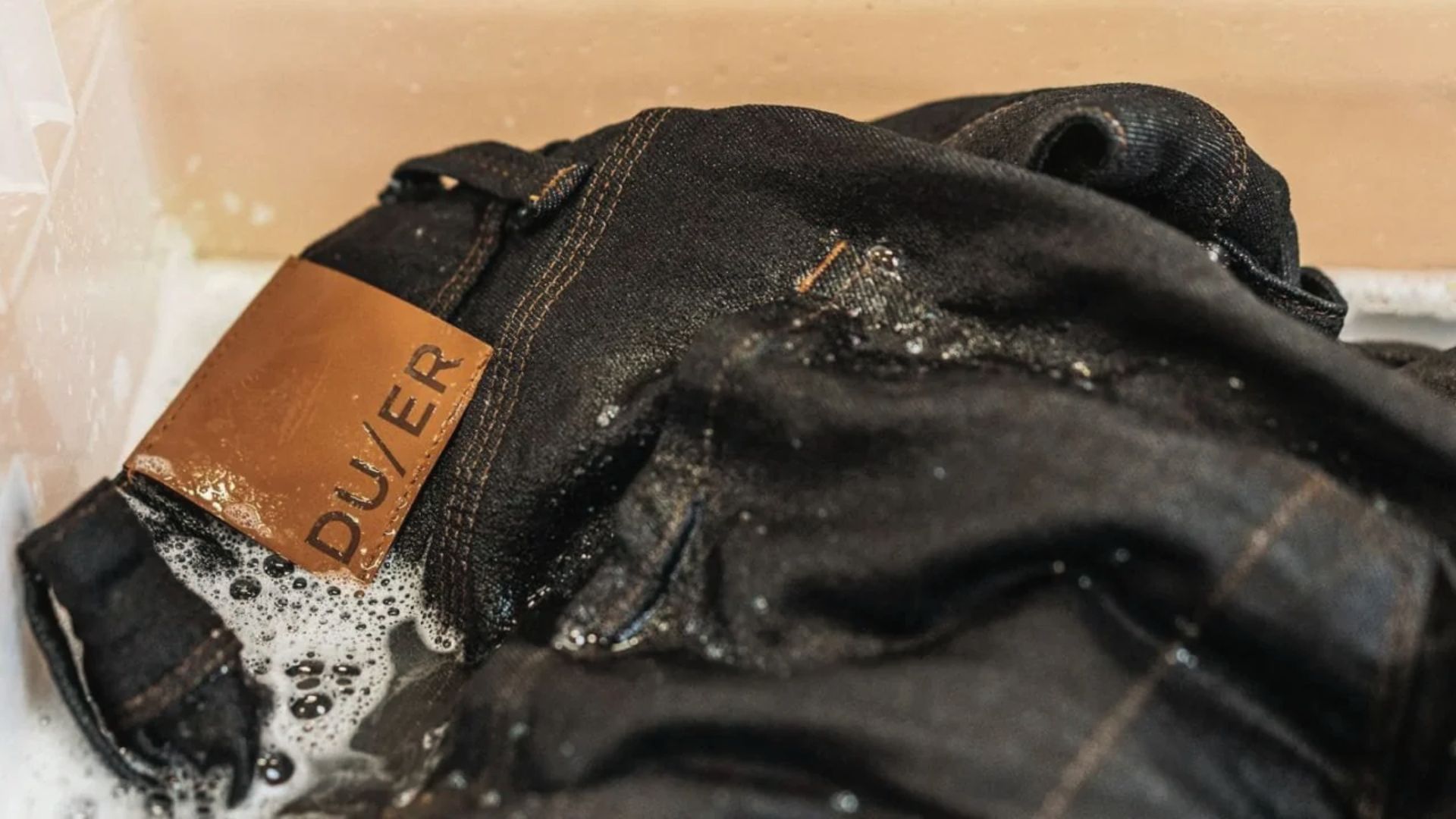 Black DU/ER brand jeans sit in a sink with soap suds clinging to the fabric.