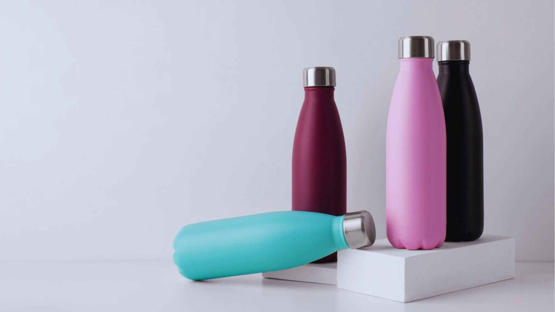 Four matte stainless steel water bottles in teal, burgundy, pink, and black with silver caps arranged on white platforms.