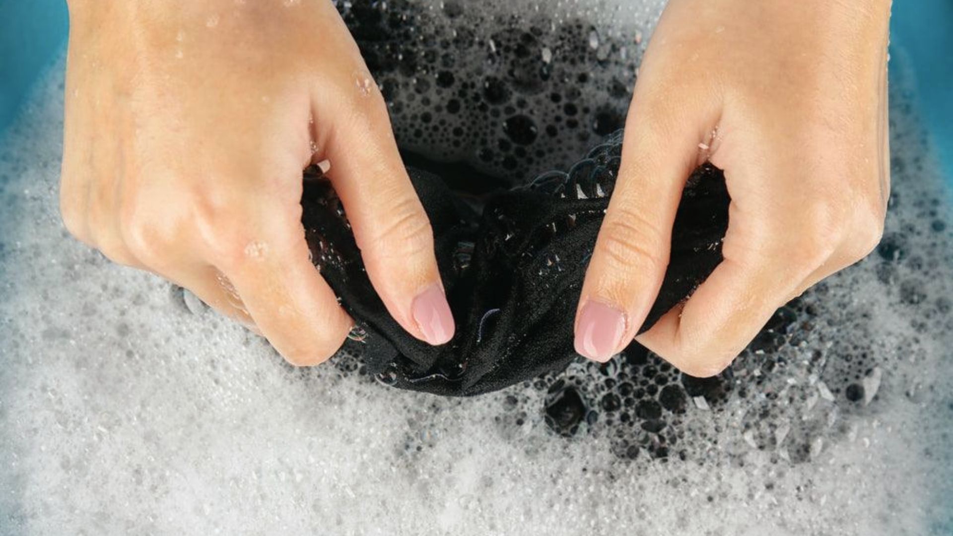 Hands wash black fabric in a blue basin with soap suds, using a gentle squeezing motion.