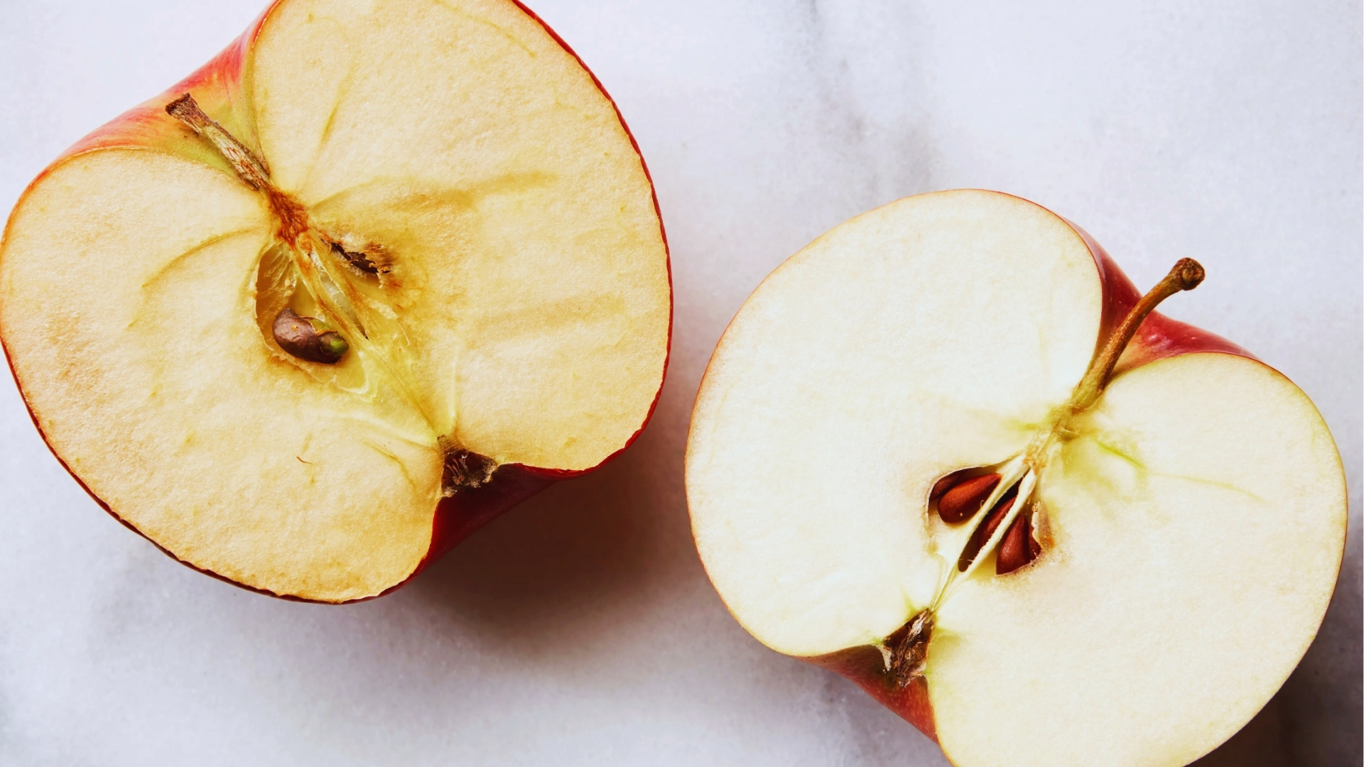 Two red apple halves cut open showing one slightly browned side and one fresh white side on the marble surface.