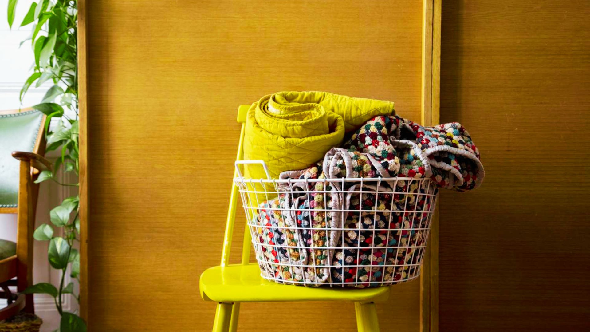 A white wire basket on a yellow chair overflowing with colorful folded blankets and quilts.
