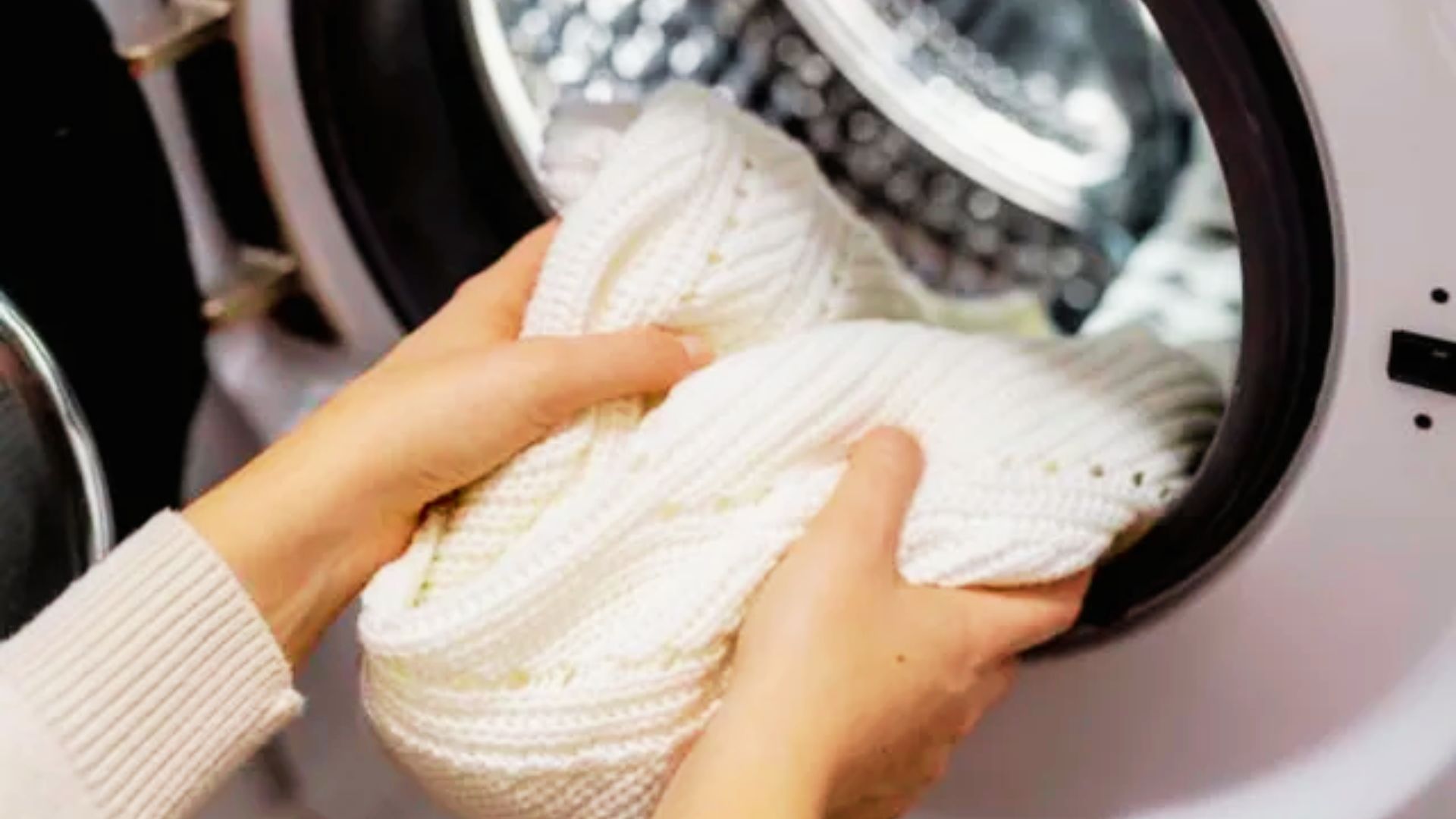 Hands loading a white knit sweater into a washing machine to disinfect using the sanitize cycle.