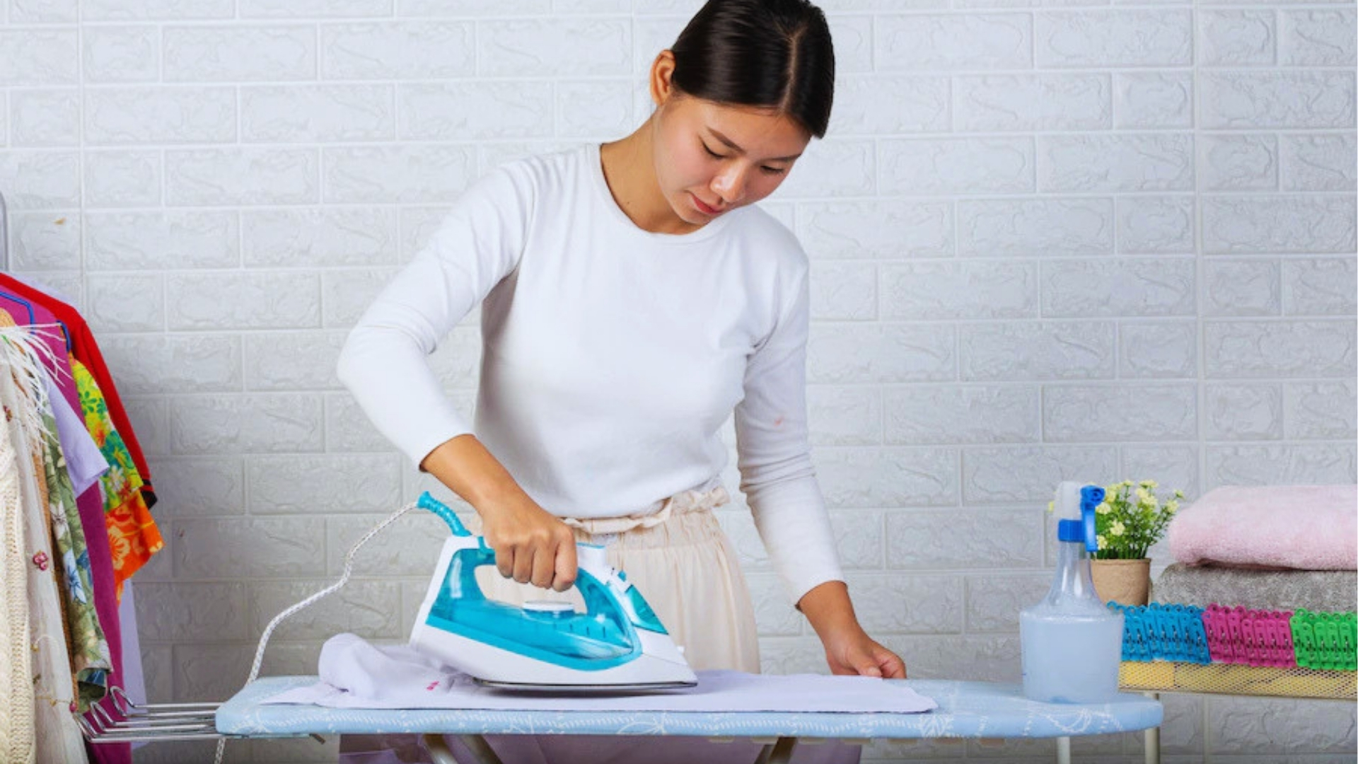 Woman in white shirt ironing white fabric on ironing board with blue steam iron, spray bottle, and folded towels nearby.