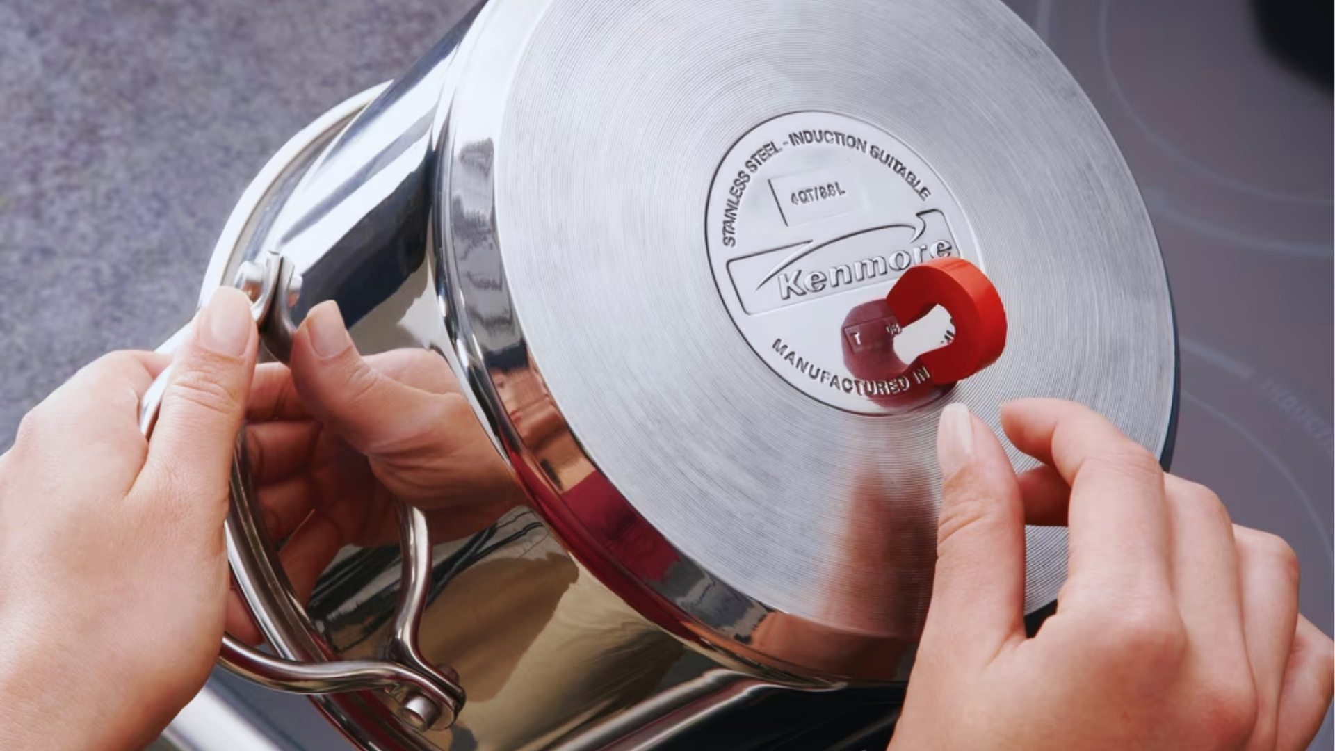 Hands testing induction compatibility of stainless steel Kenmore pot base with red horseshoe magnet over induction cooktop.