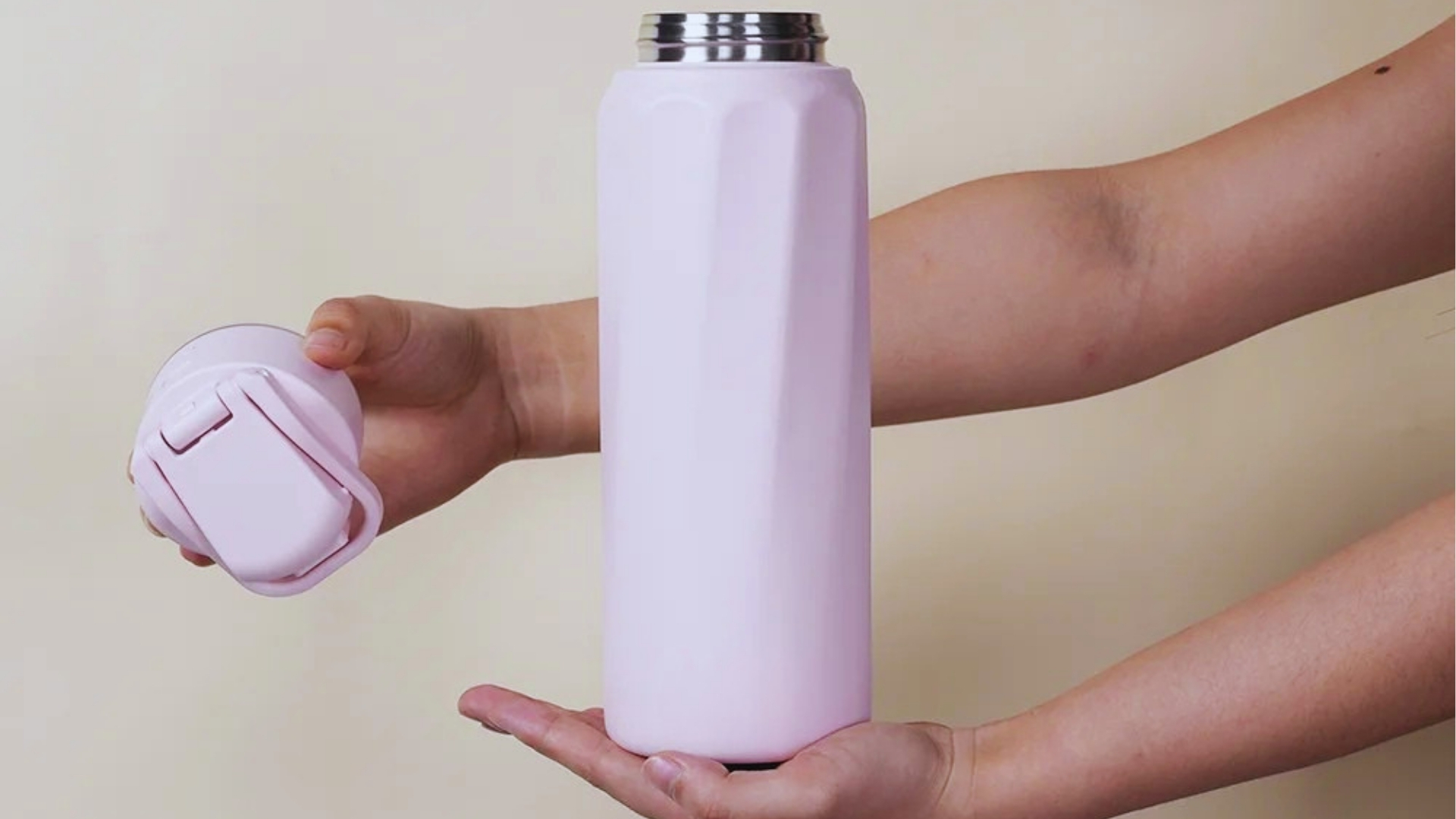 Hands holding pale pink stainless steel water bottle with geometric design and matching flip-top lid removed against beige background.