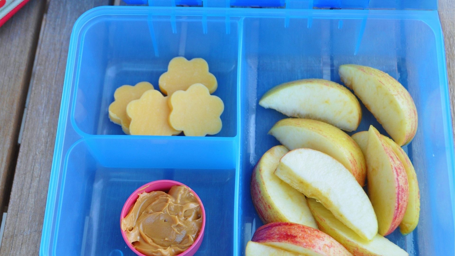 Blue divided lunch container holding fresh apple slices, flower-shaped cheese pieces, and small pink cup of peanut butter on wooden table.