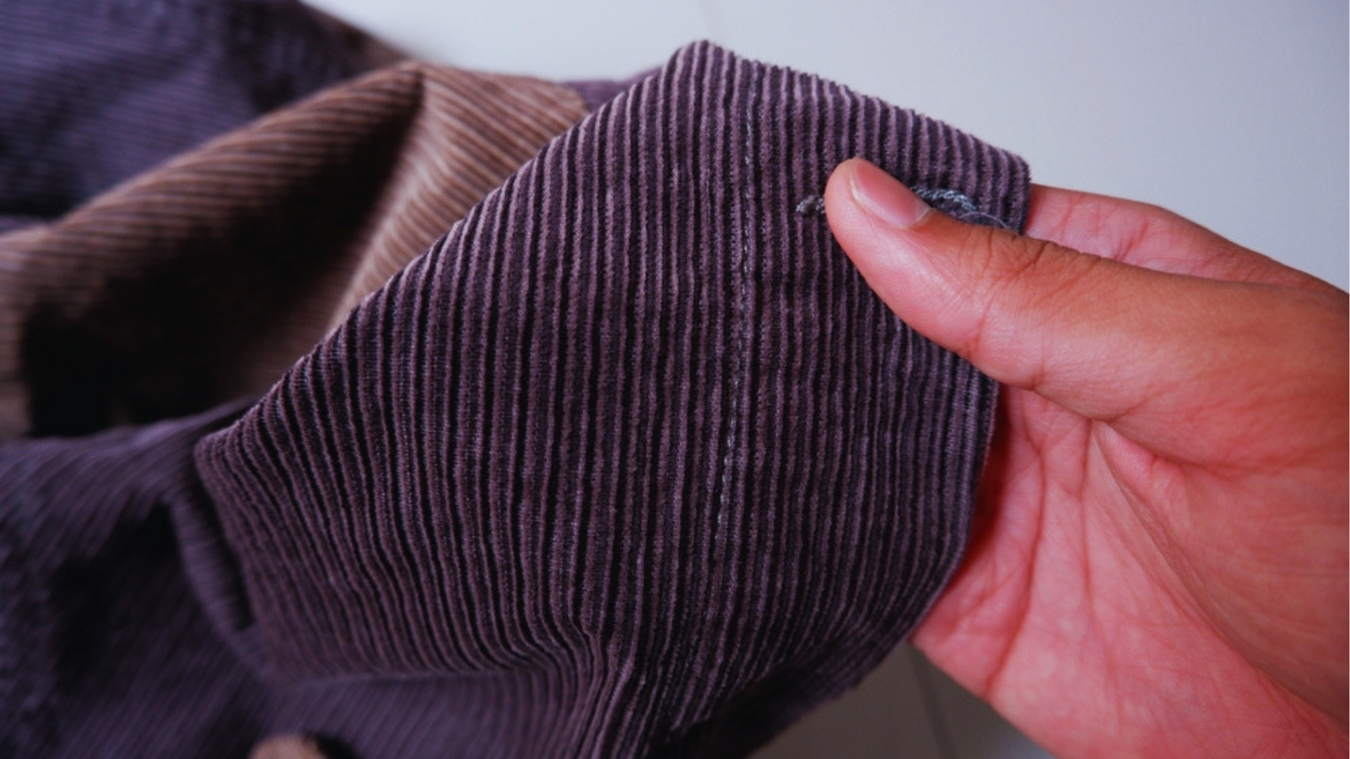 Close-up of hand holding folded plum and brown corduroy fabric showing raised ridged wale texture.