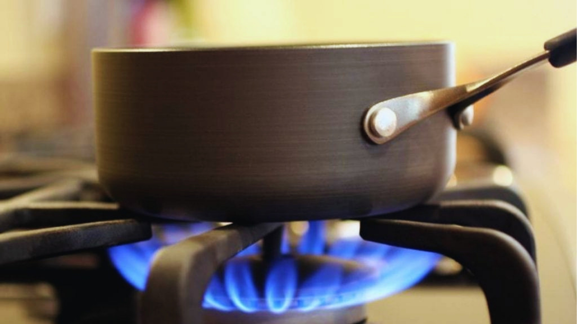Dark non-stick saucepan with handle on gas stove burner with visible blue flames underneath against blurred kitchen background.