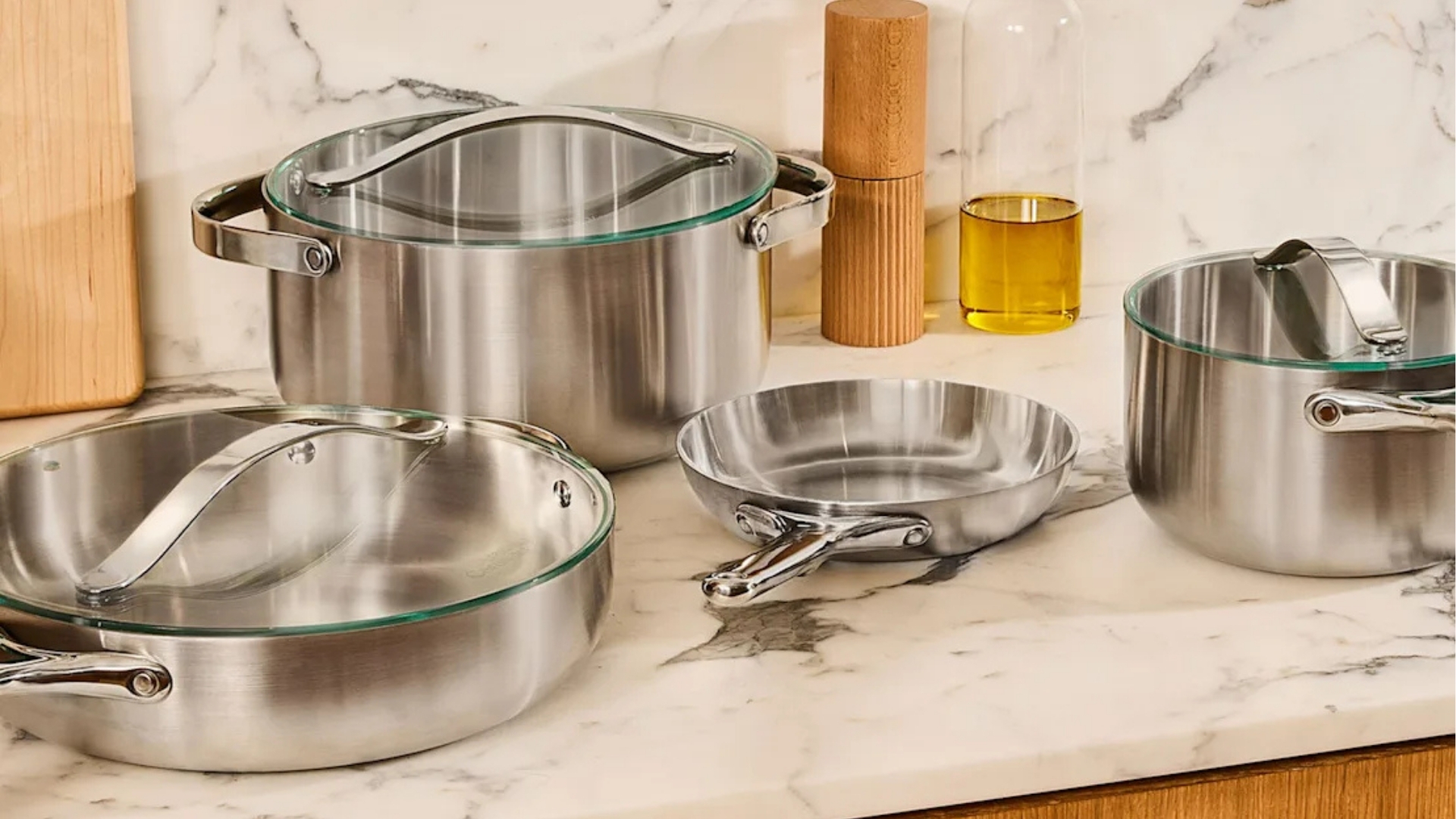 Set of polished Clad Cookware with glass lids including pots and frying pan arranged on marble countertop.