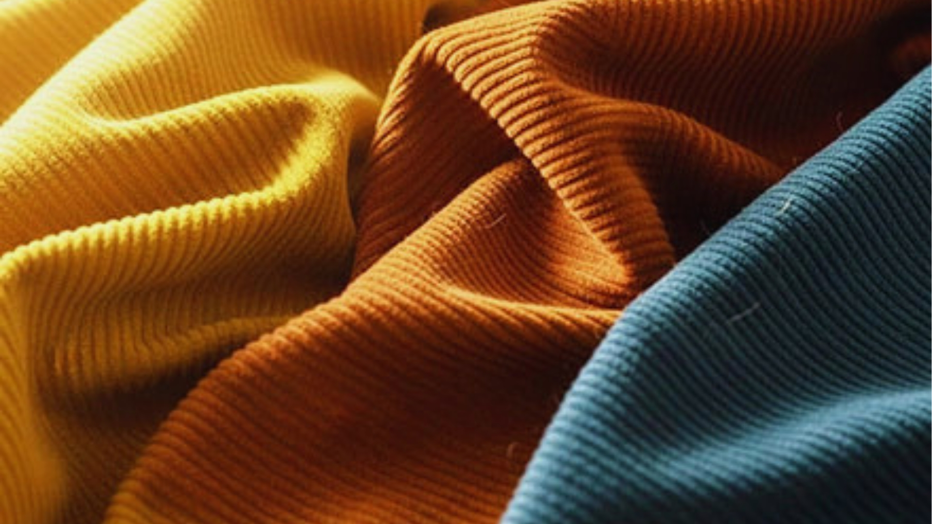 Folded yellow, rust orange, and teal corduroy fabrics with soft ridged texture in warm sunlight.