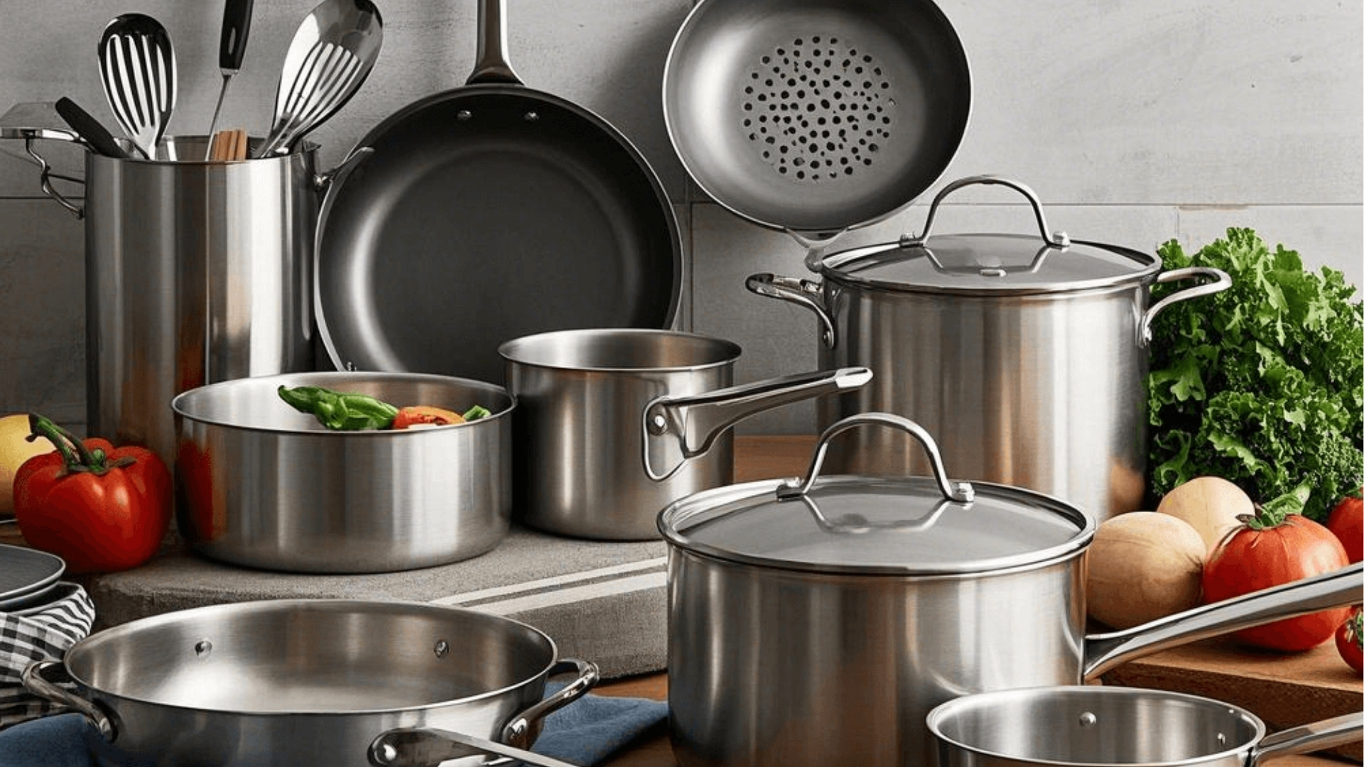 Assorted stainless steel cookware set with pots, pans, and utensils displayed with fresh vegetables on kitchen counter.