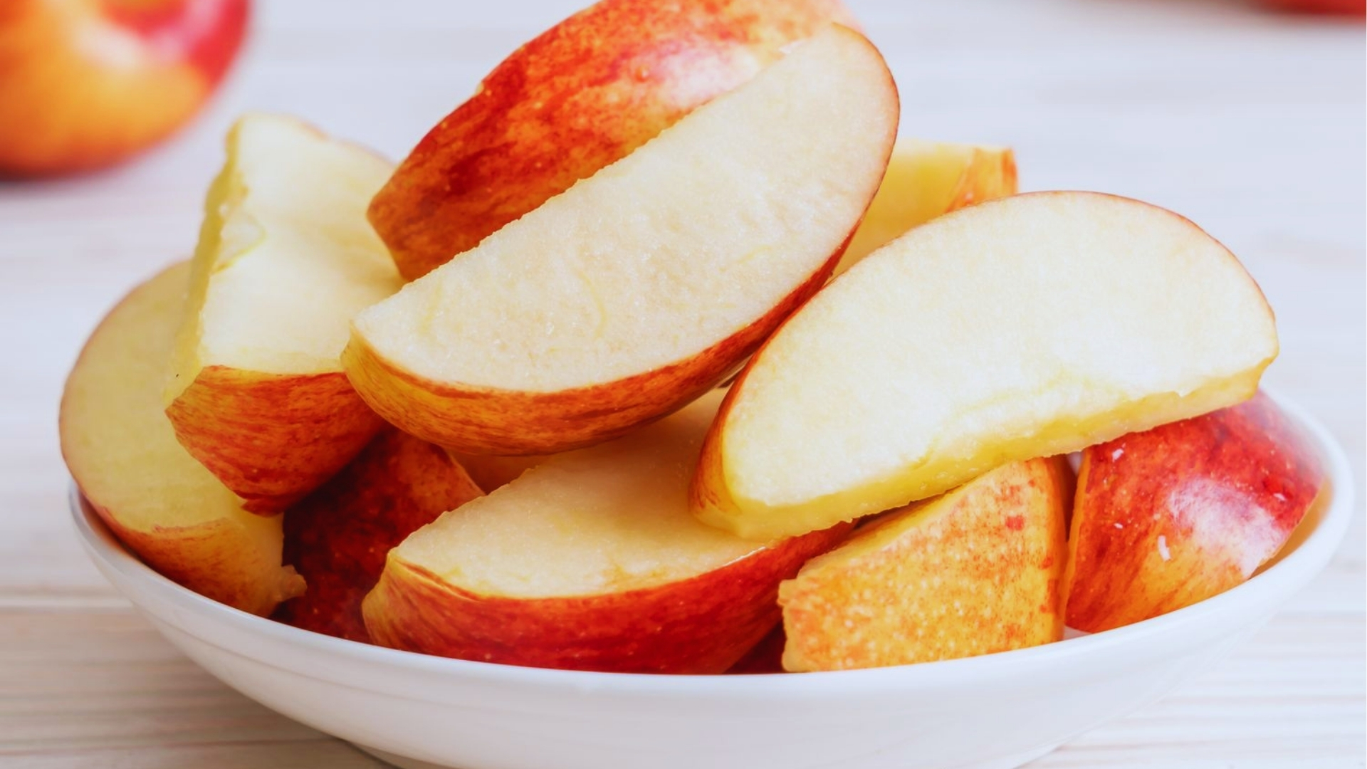 Close-up of fresh red apple slices piled in white ceramic bowl showing crisp white flesh against a light wood background.