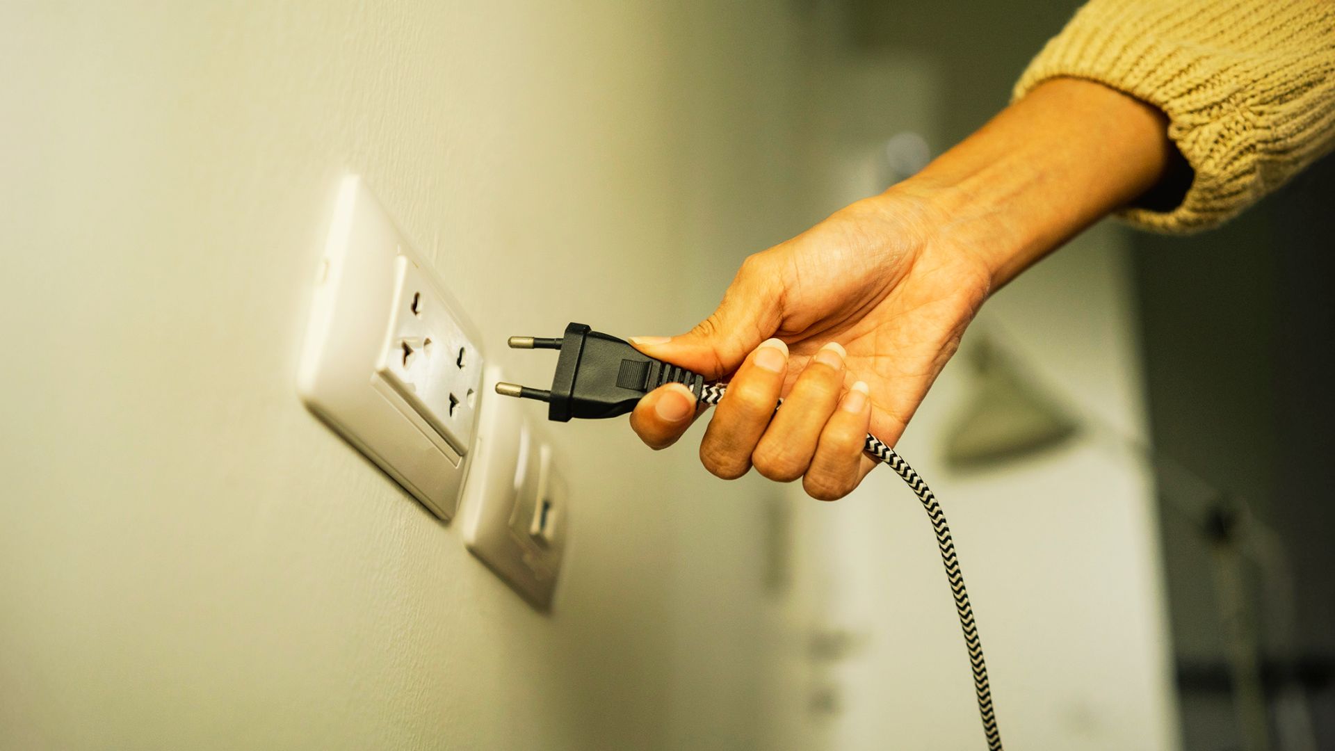 Hand unplugging a black power cord from a wall outlet to stop standby power consumption.