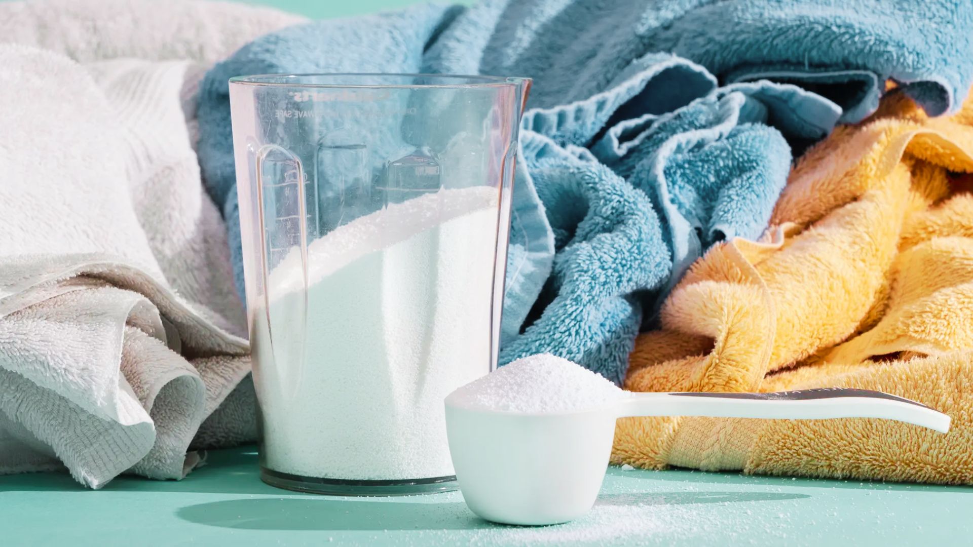 A measuring cup with borax in it and a scoop of laundry detergent sit in front of colorful, clean towels.