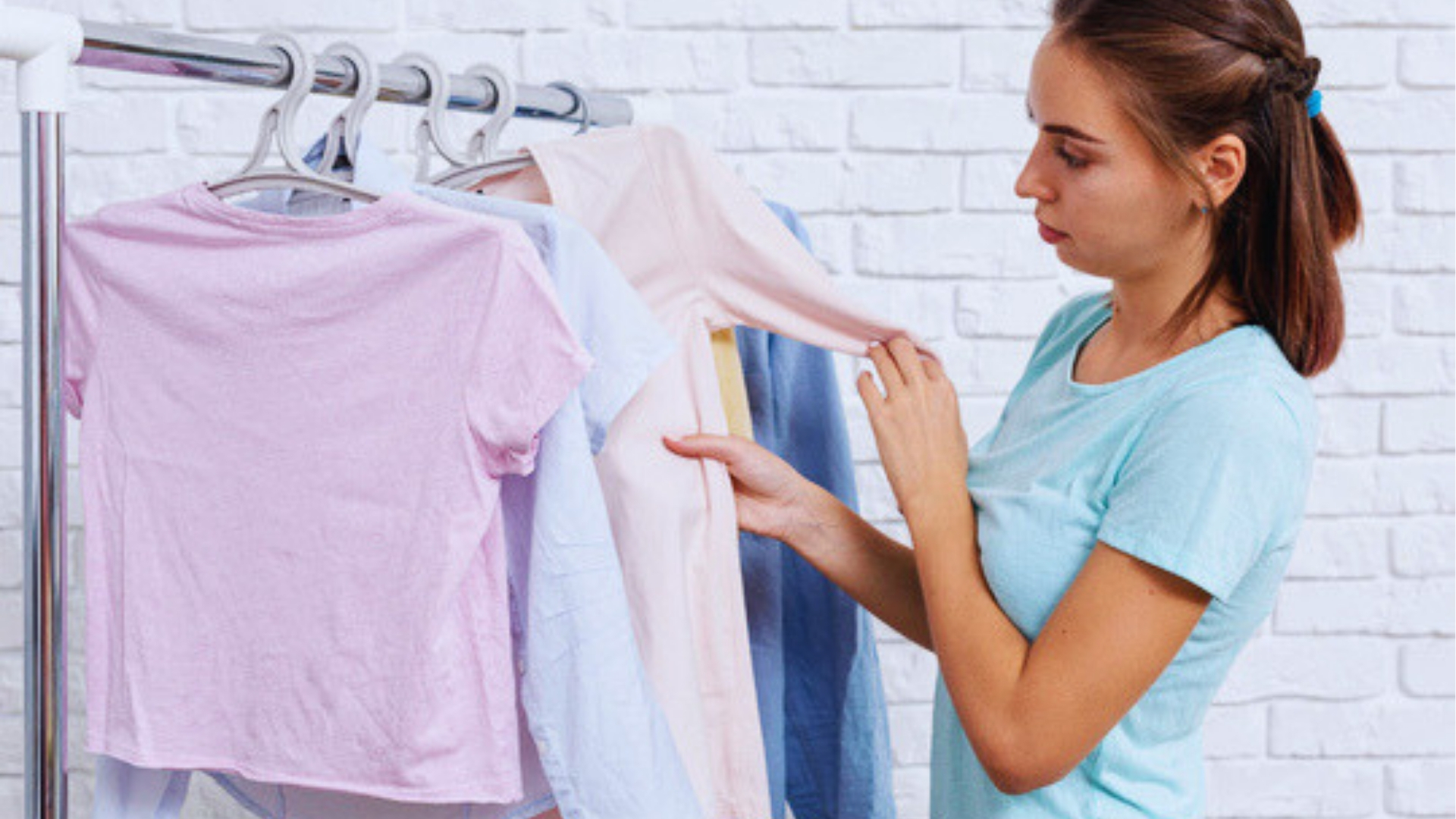 Young woman in teal shirt examining sleeve of pastel shirt hanging on clothing rack against white brick wall.