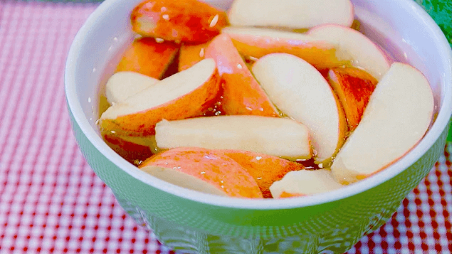 Red apple slices soaking in water inside a green ceramic bowl on red gingham cloth to prevent browning.