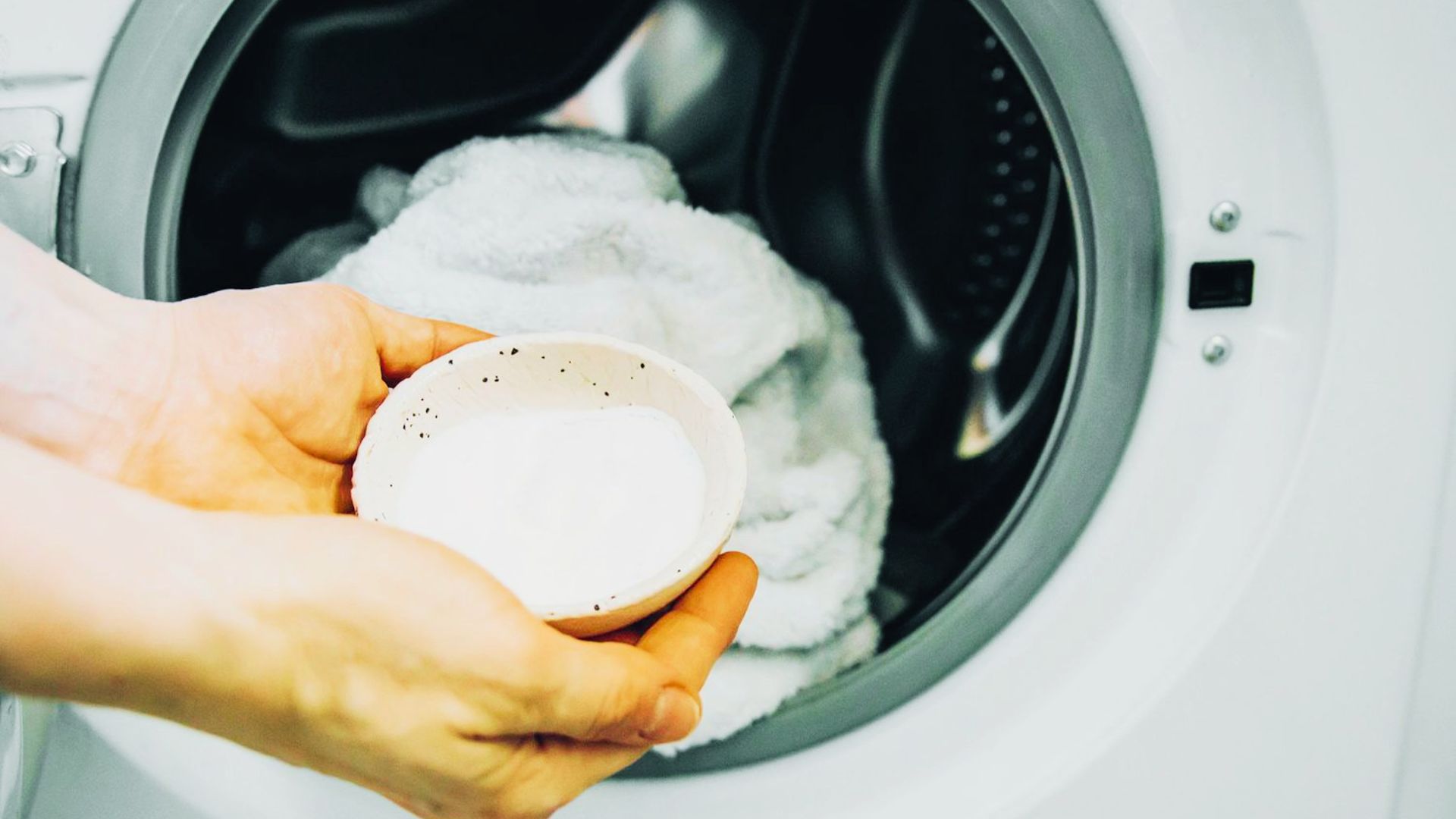 Hands hold a small bowl of baking soda over a washing machine filled with white towels.