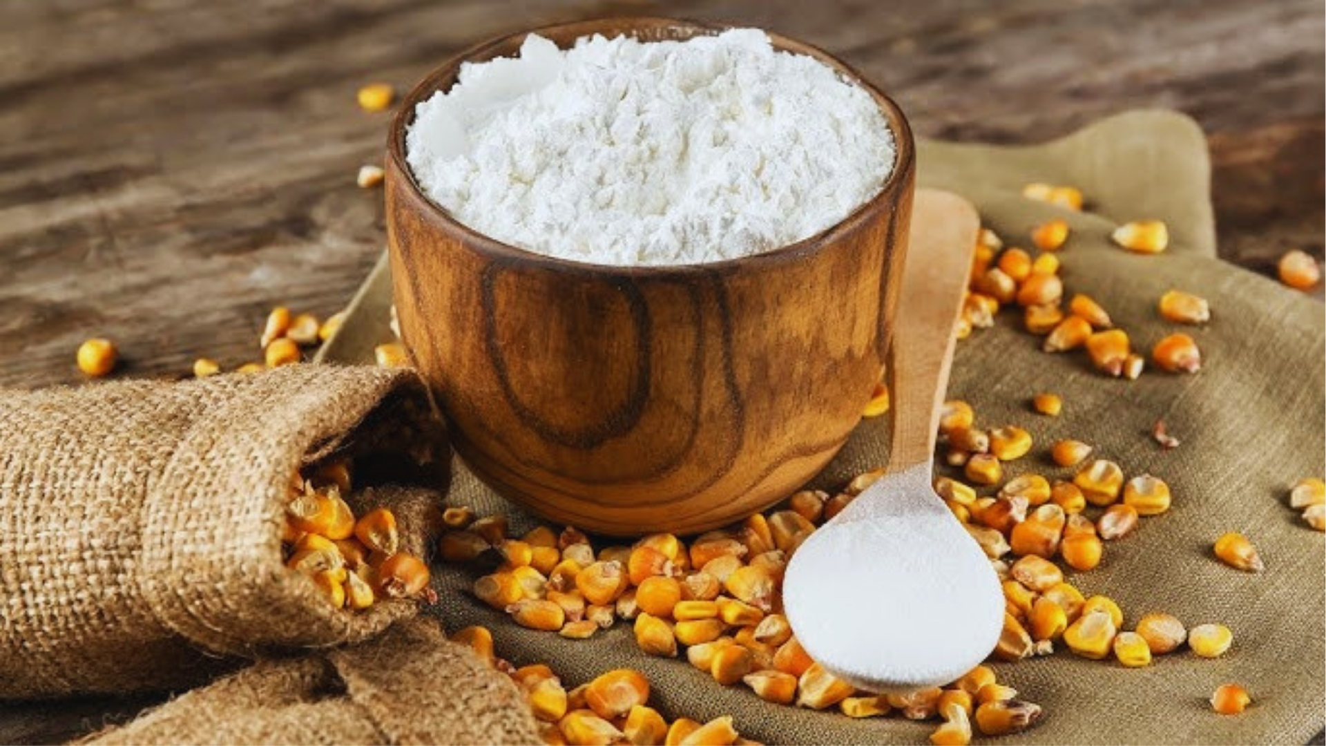 Wooden bowl filled with white starch powder on burlap, with wooden spoon and scattered corn kernels.