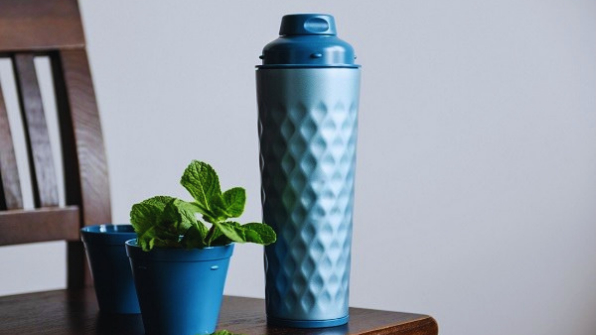 Blue textured stainless steel water bottle with flip lid on wooden table next to small potted mint plant and blue cup