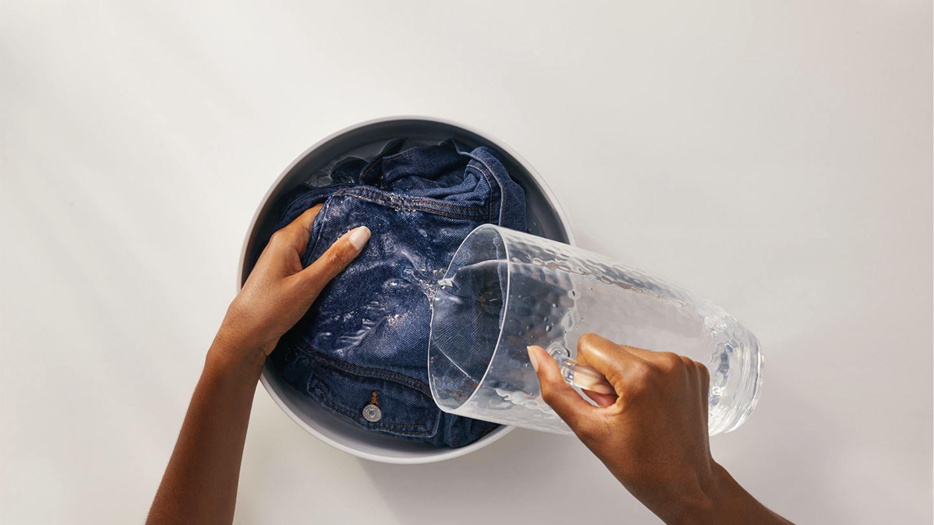 Hands pour water over jeans in a bowl to pre-treat ink stains before washing stained clothes.