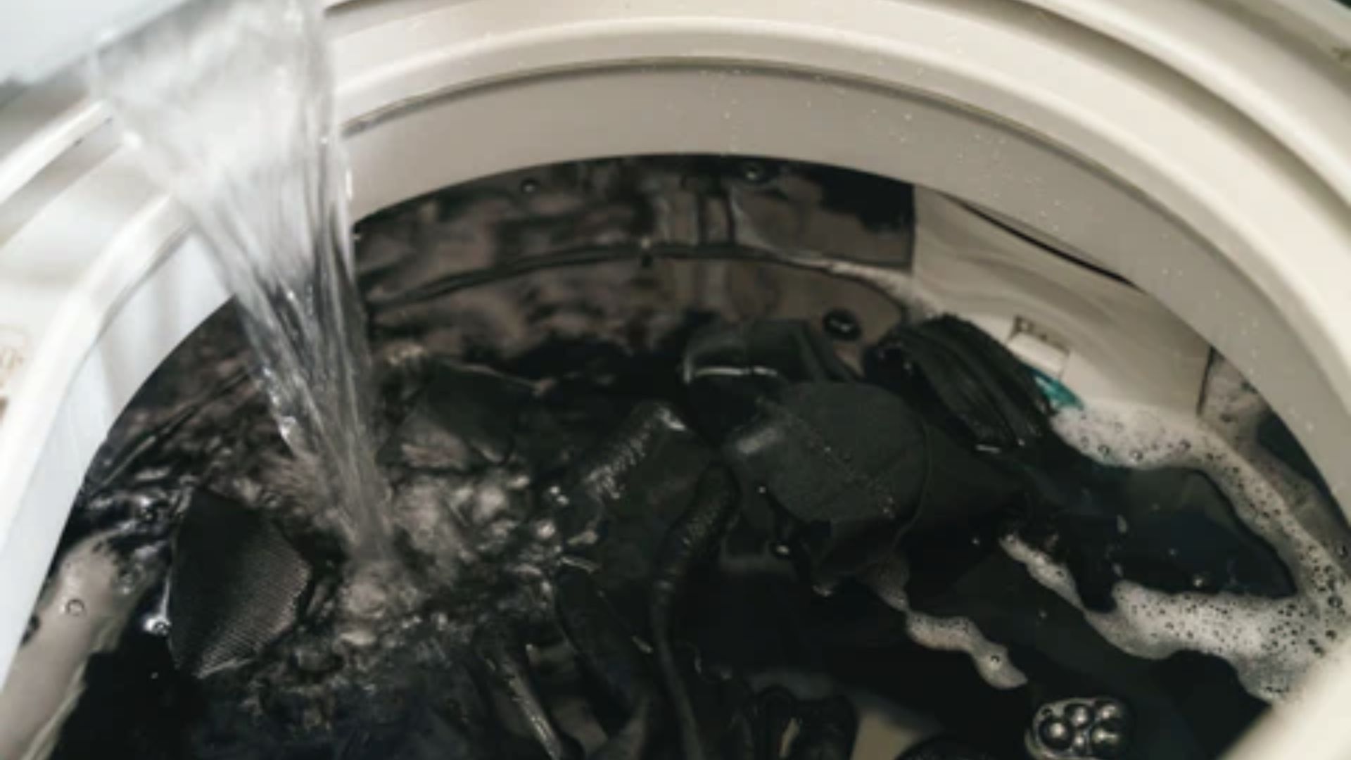 Water pours into a top-load washing machine filled with black jeans and suds.