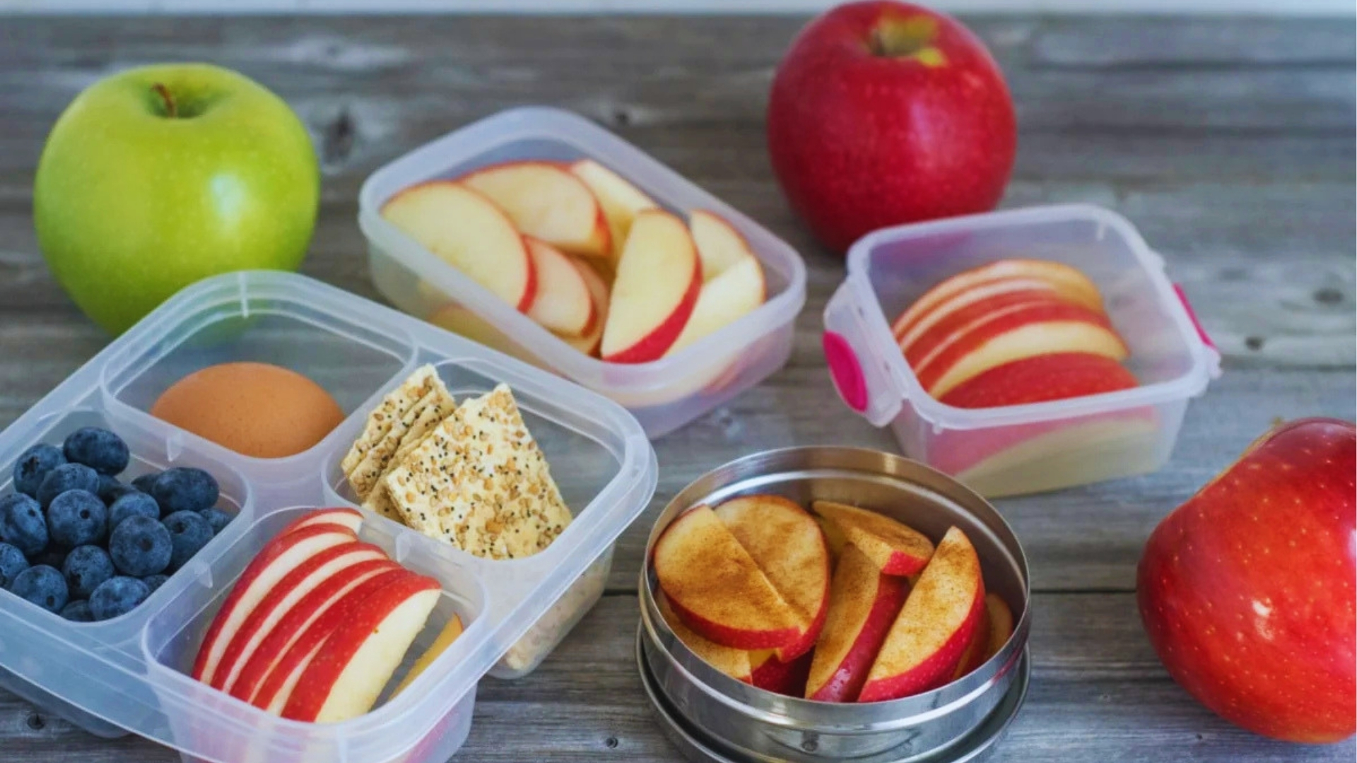 Various lunch containers with sliced red apples, blueberries, boiled egg, crackers, and cinnamon apple slices next to whole green and red apples on a wood surface.